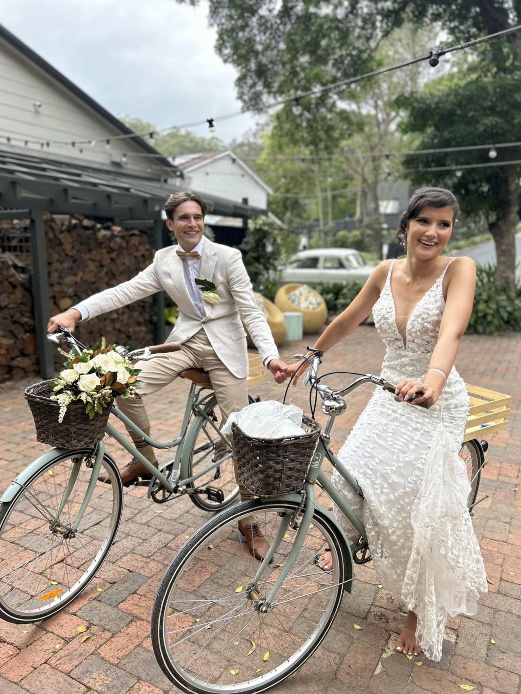 Smiling bride and groom ride vintage-style bicycles together in an outdoor wedding setting.