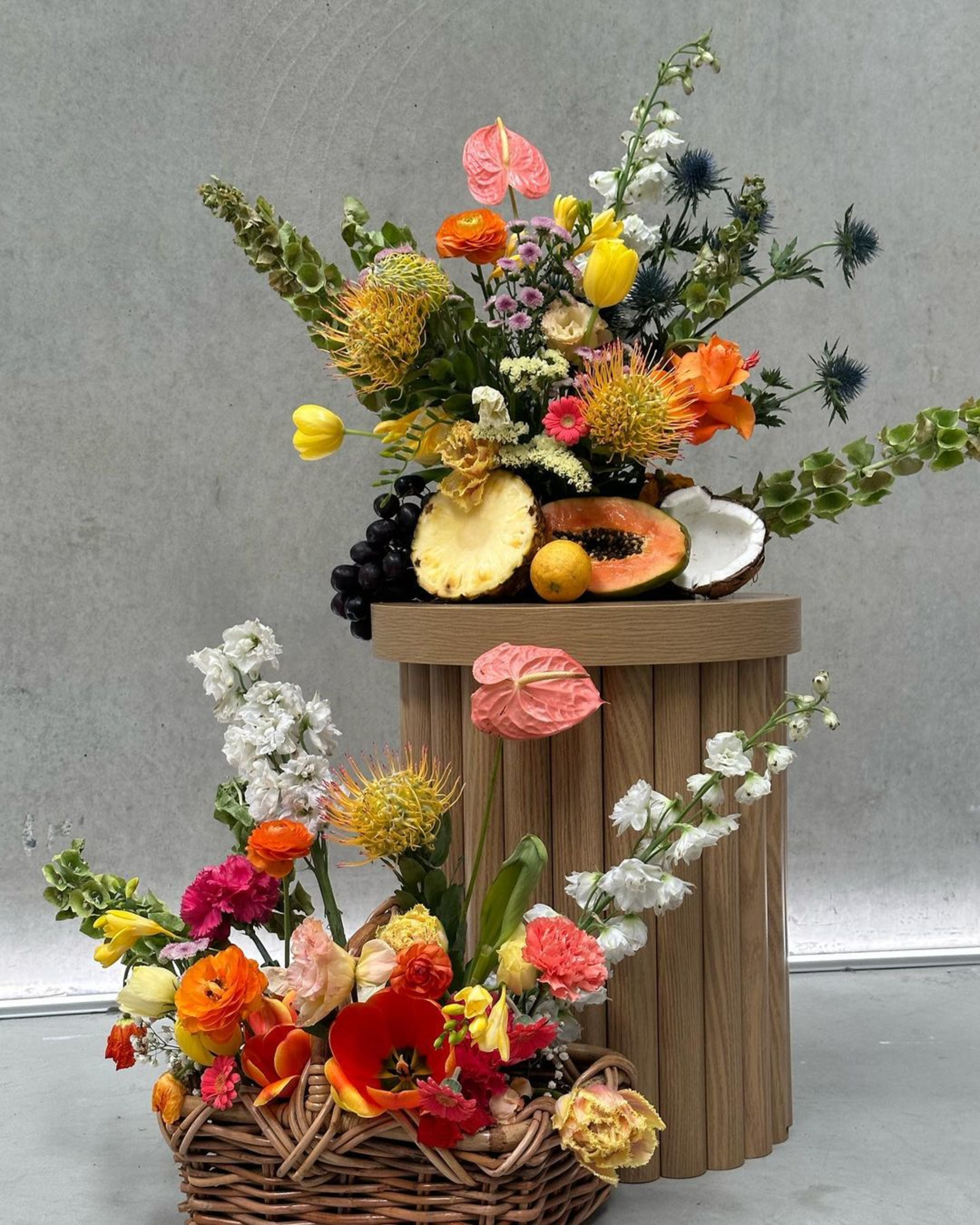 Vibrant floral arrangements with tropical fruit displayed in a basket and on a wooden pedestal against a neutral backdrop.