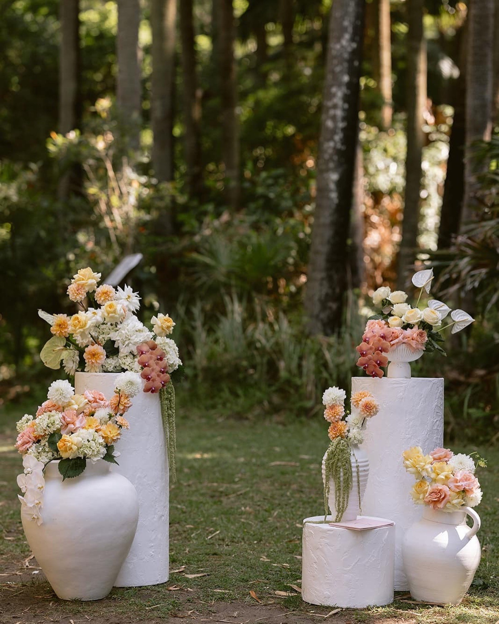 White pedestals and vases with pastel floral arrangements set in a lush forest wedding ceremony space.