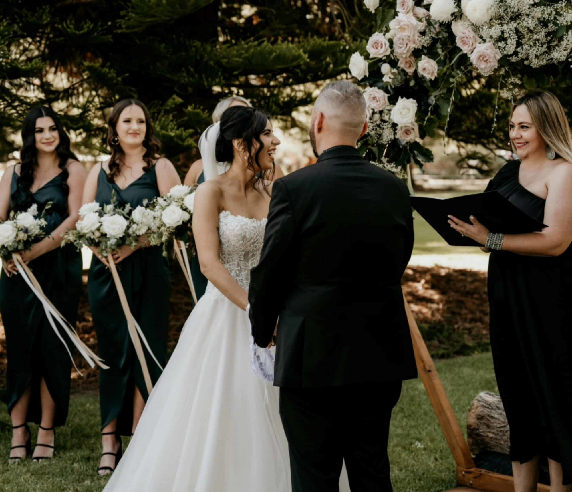 Outdoor wedding ceremony with couple exchanging vows beneath a floral arbor and bridesmaids holding white bouquets.