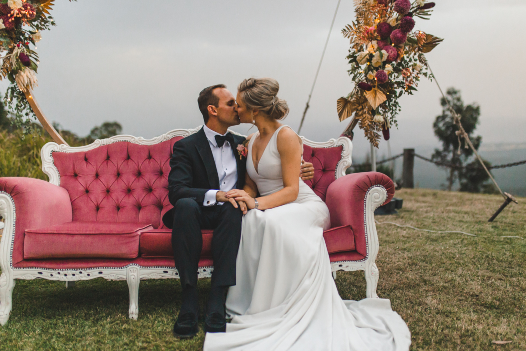 Bride and groom kiss on a pink vintage sofa with floral arrangements in an outdoor wedding setting.