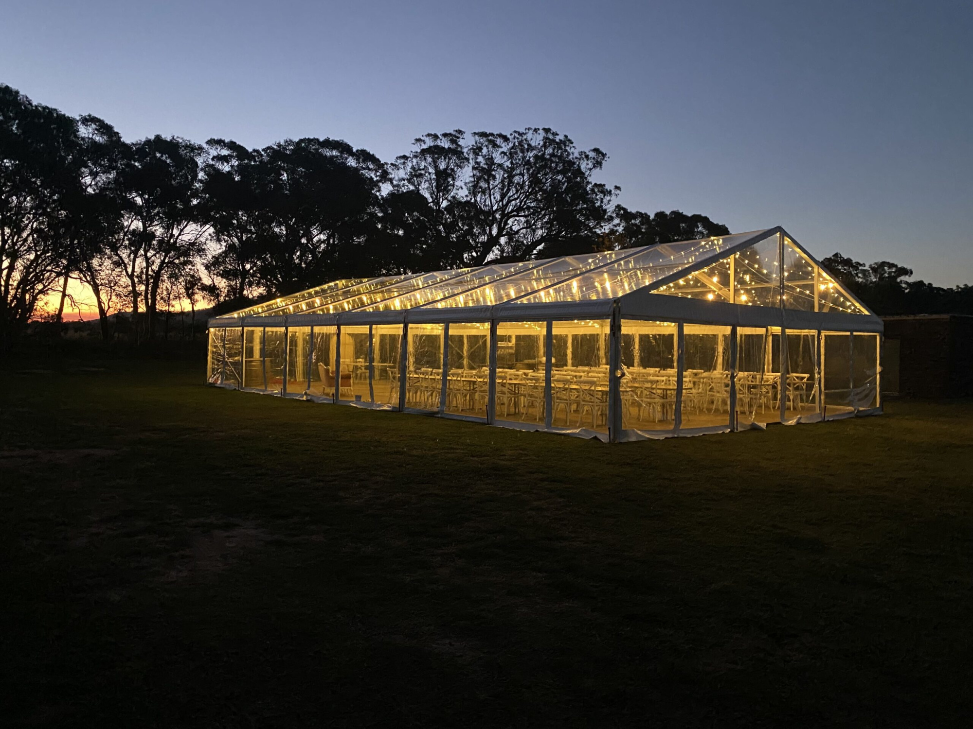 Clear marquee wedding reception tent glowing with string lights at dusk in an outdoor field.
