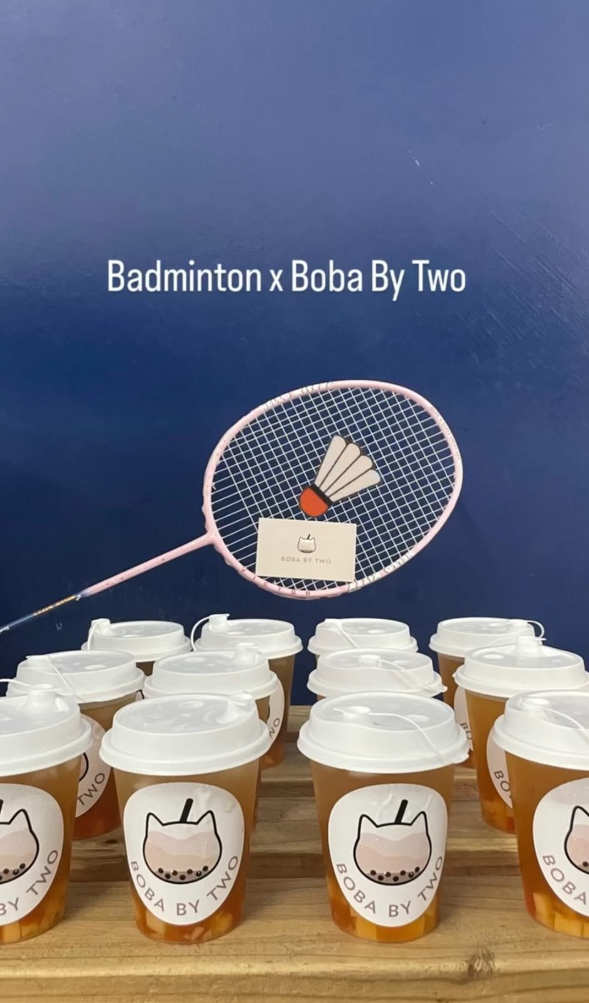 Multiple Boba By Two drinks arranged on a wooden surface with a pink badminton racket against a blue background.