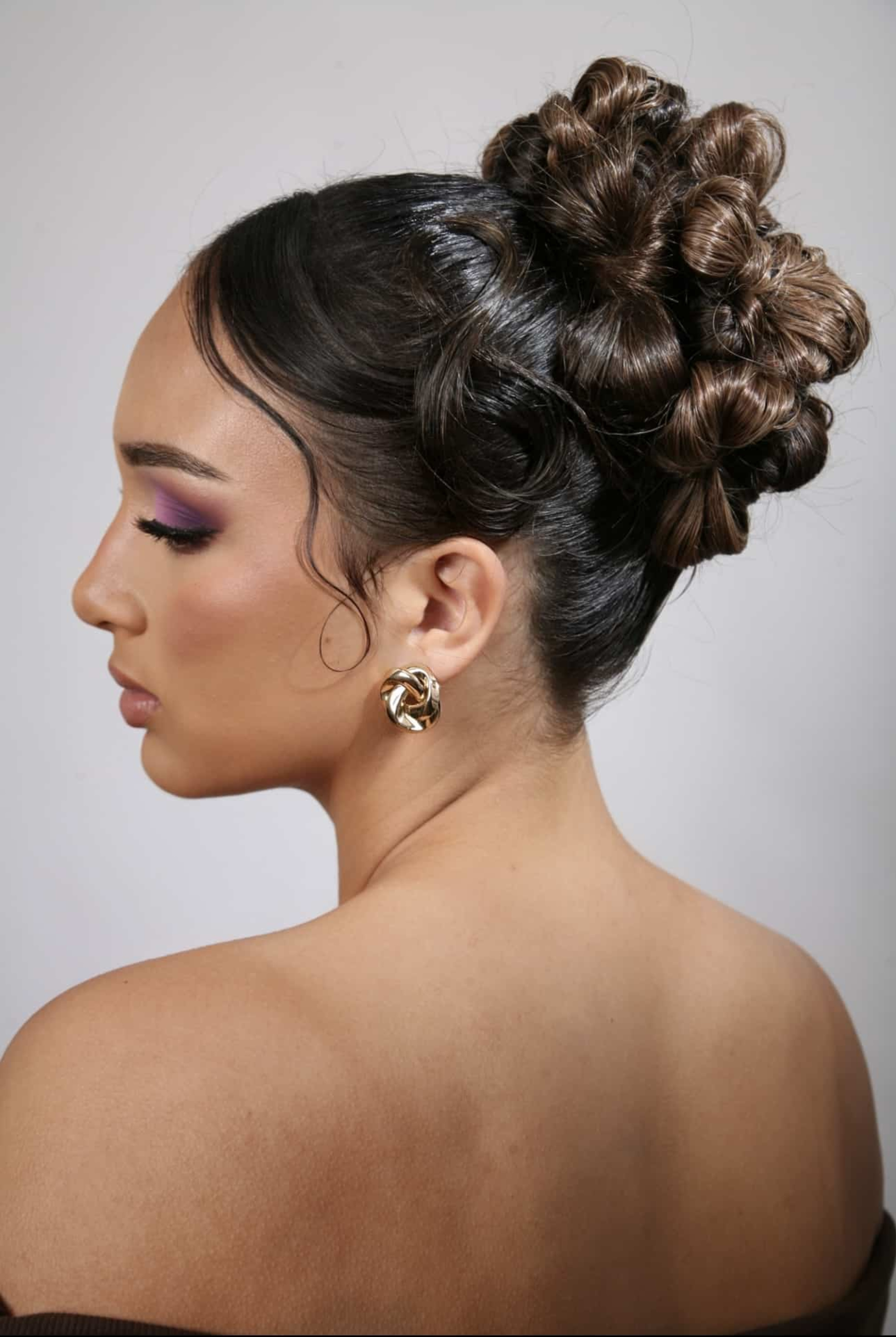 Profile of a woman with an intricate curled bridal updo and glam makeup against a plain background.