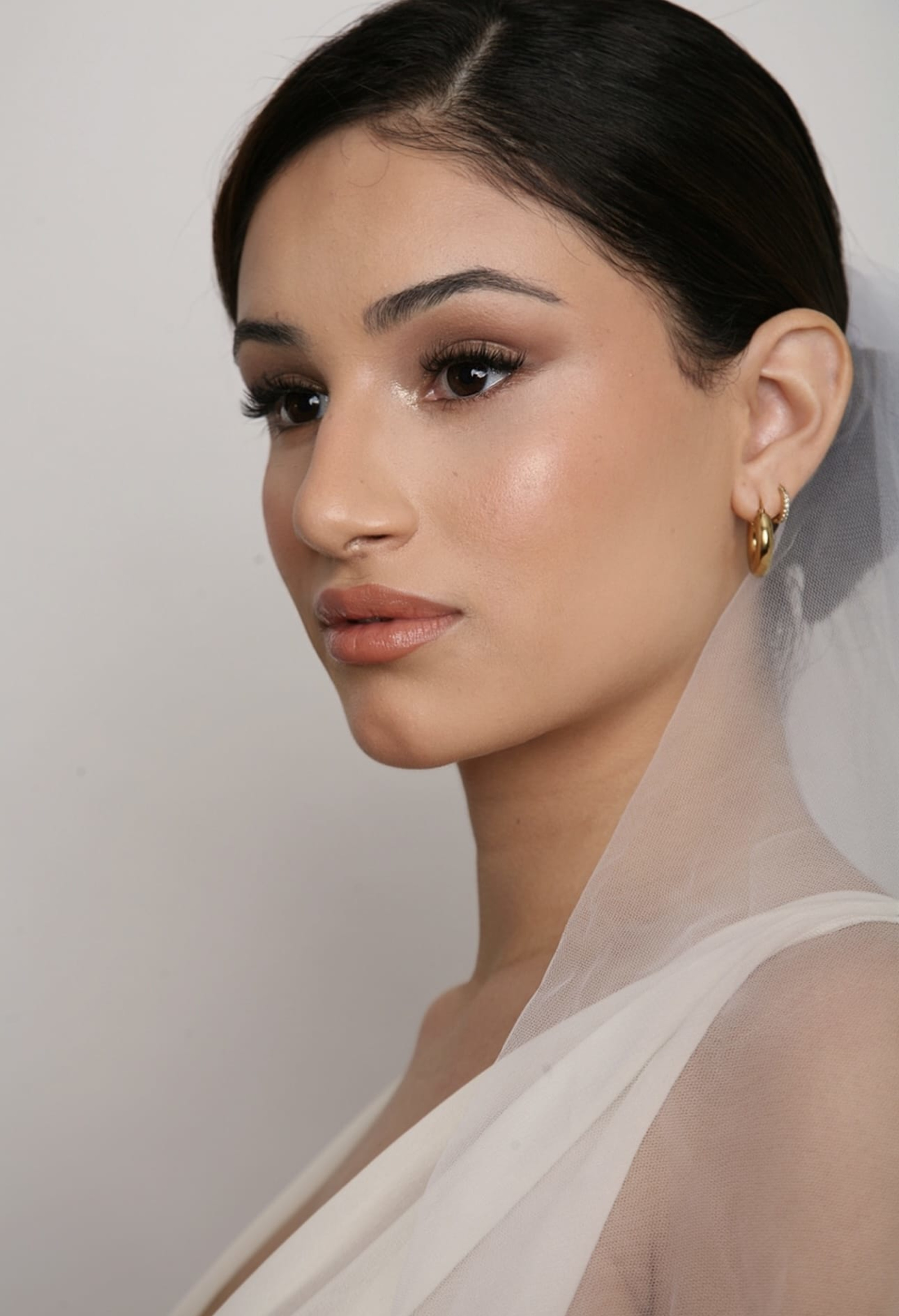 Close-up bridal portrait with natural glam makeup, veil, and gold hoop earrings against a neutral background.