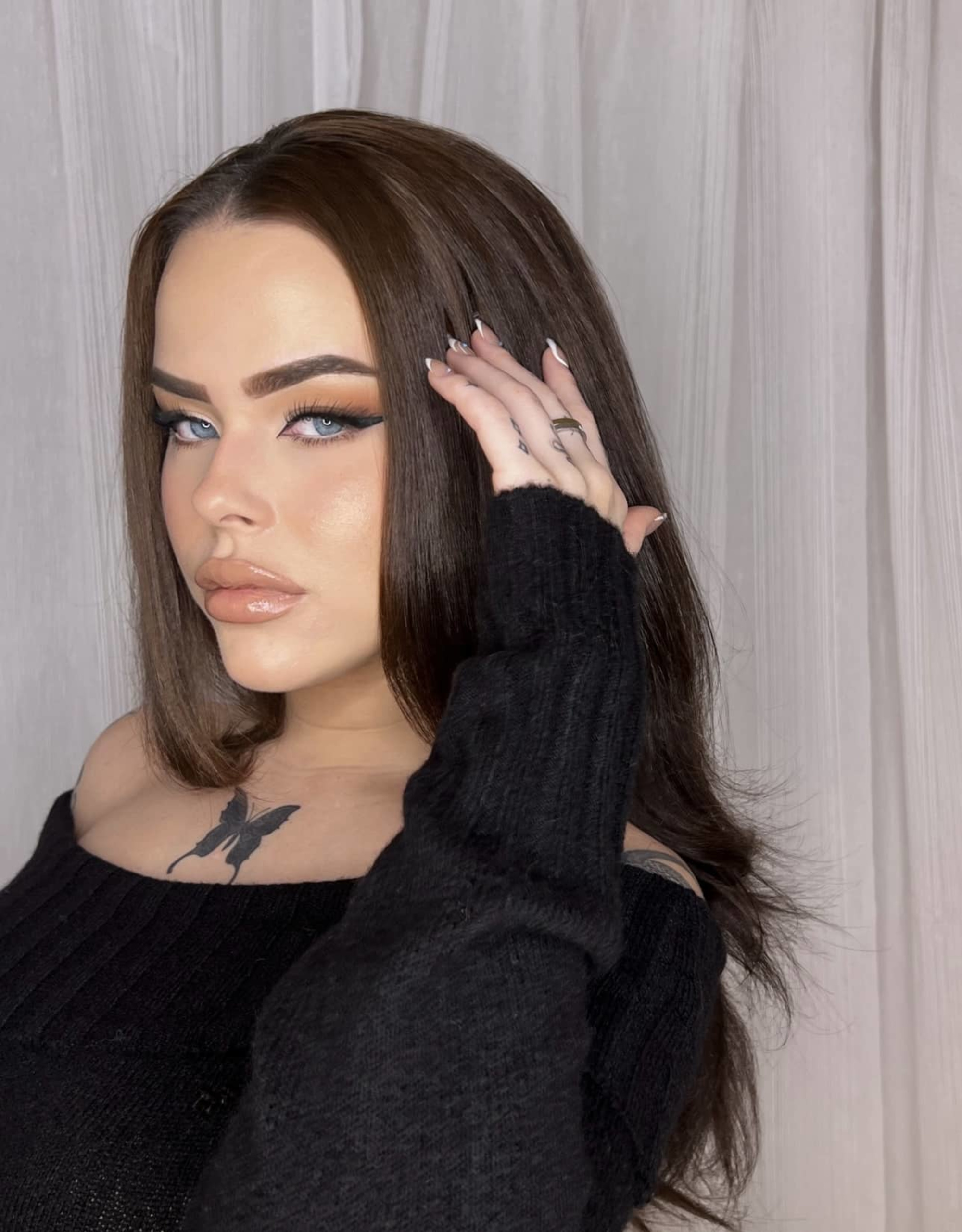 Close-up portrait of a woman with glam makeup, long brown hair, and a black top against a light curtain backdrop.