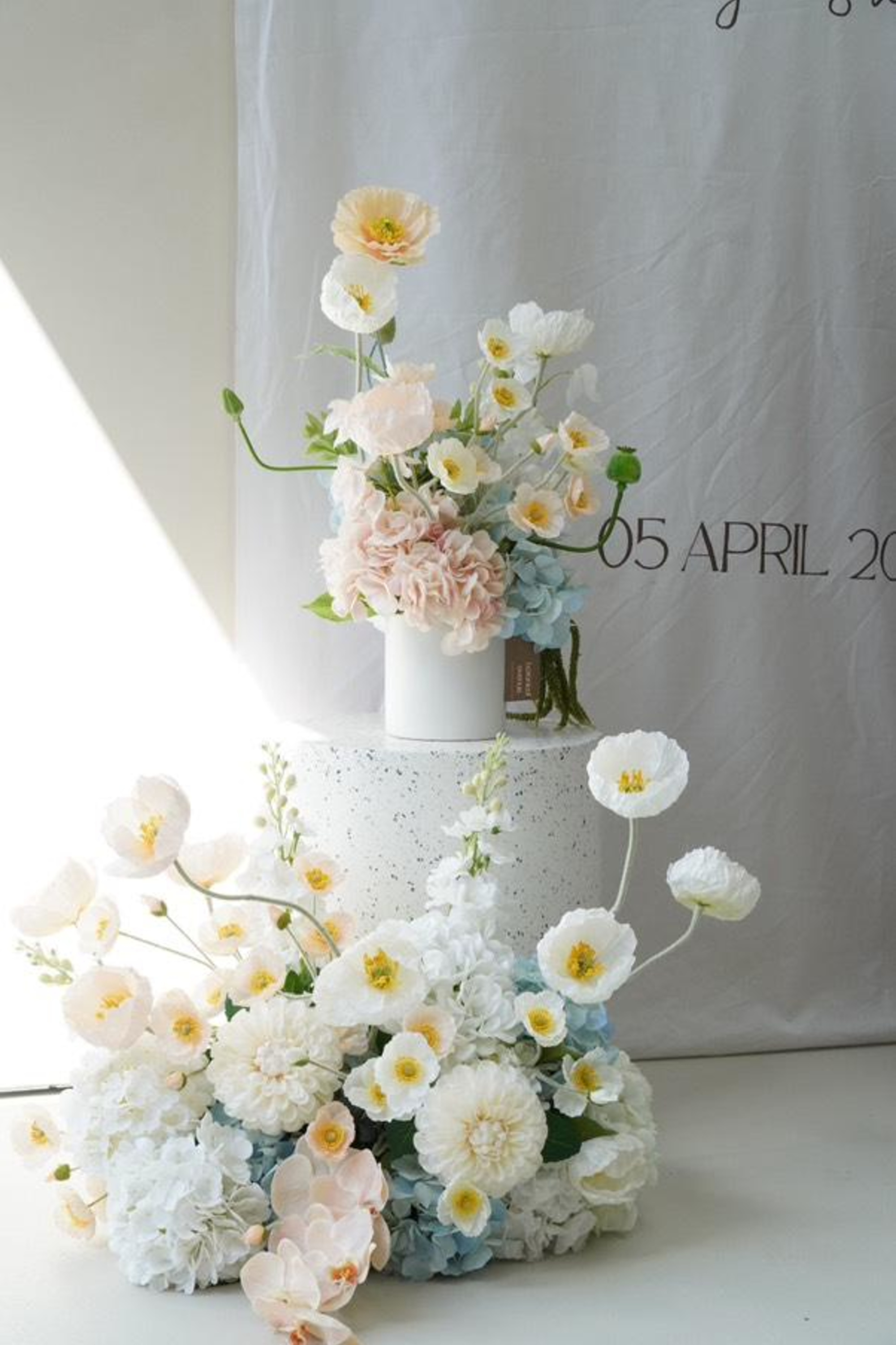 Pastel white and blush floral arrangements styled on pedestals against a soft backdrop with wedding date text.