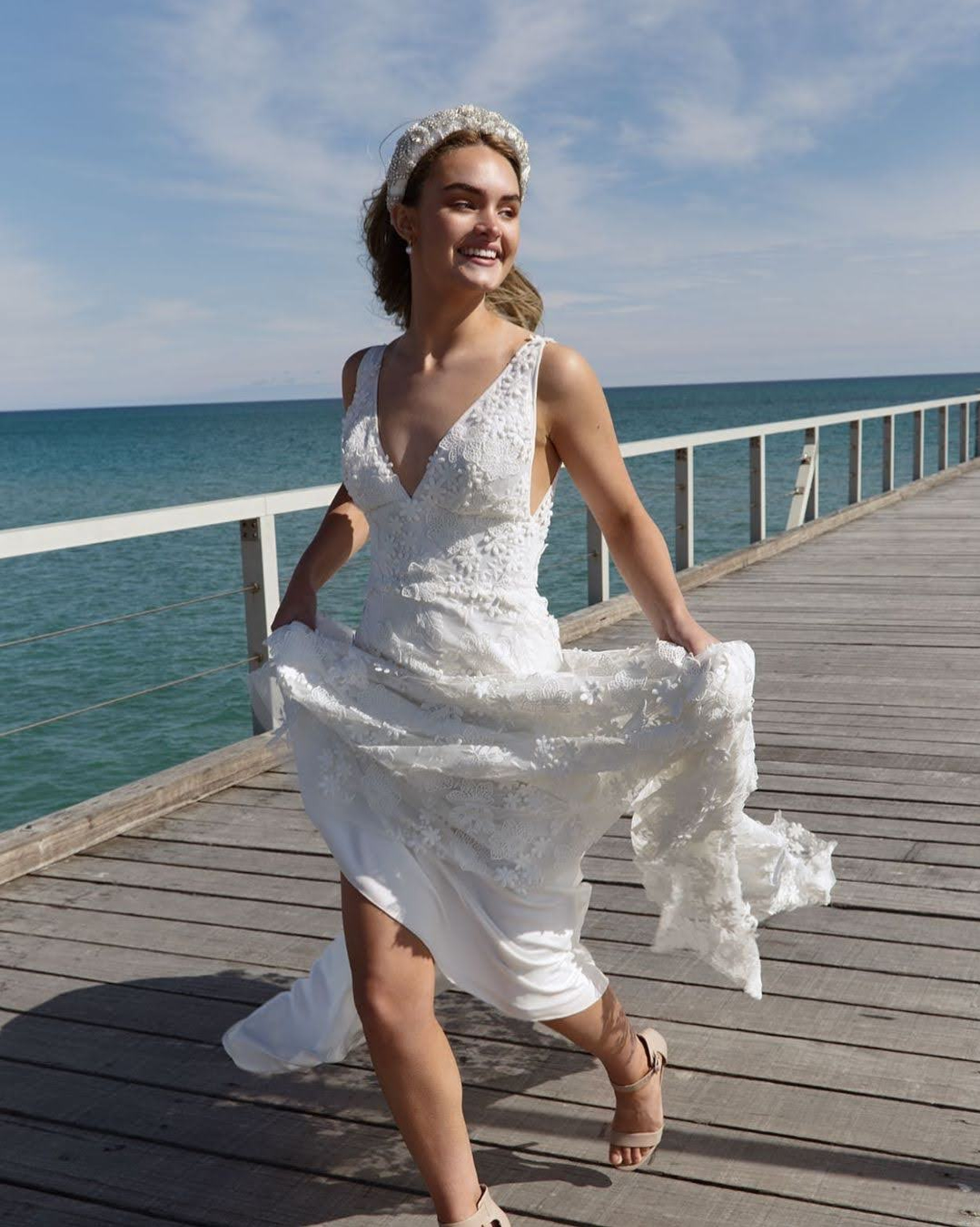 Smiling bride runs along a wooden pier in a lace wedding dress by the ocean.