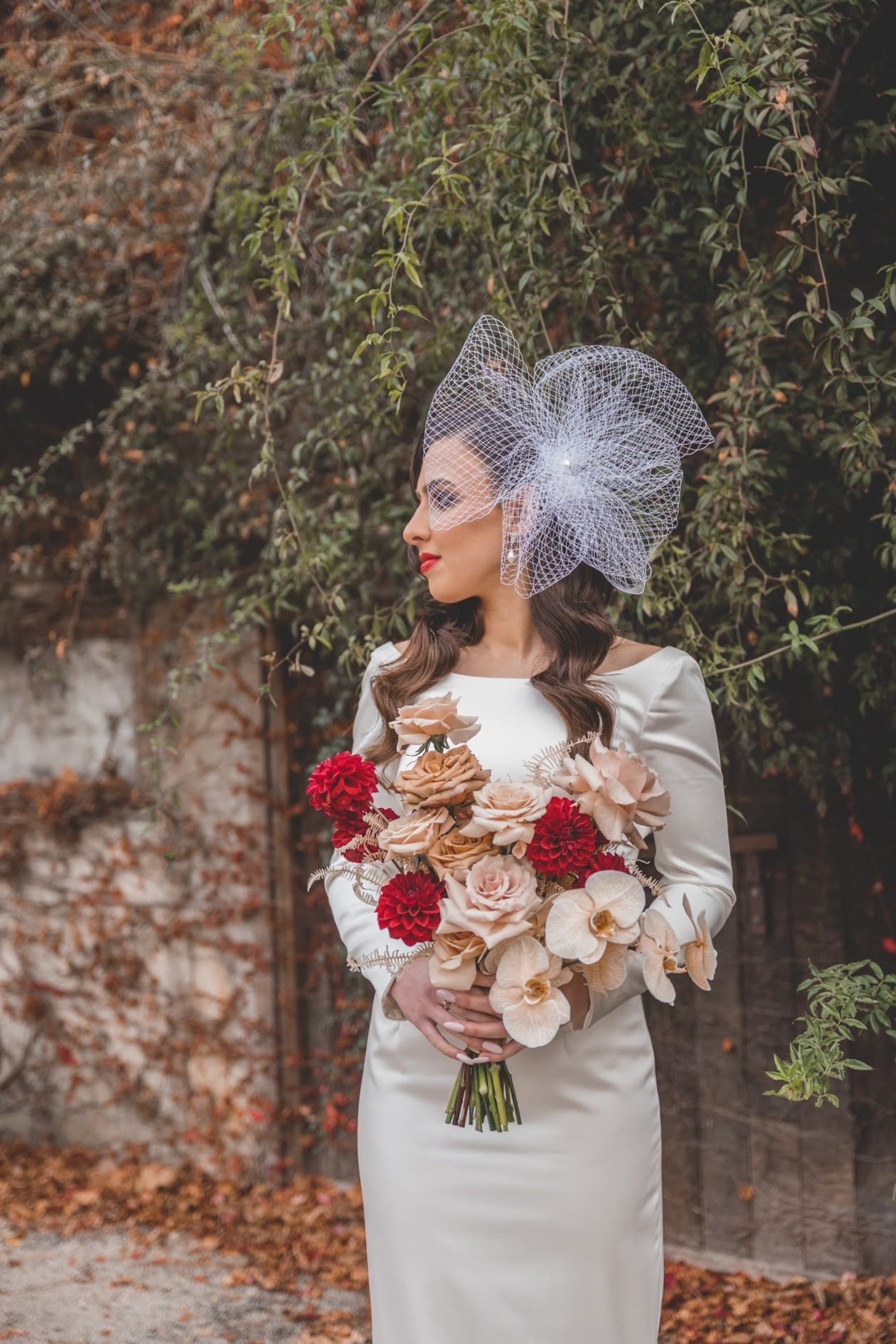 Bride in an ivory gown holding a red and neutral bouquet, wearing a dramatic netted headpiece against leafy outdoor backdrop.
