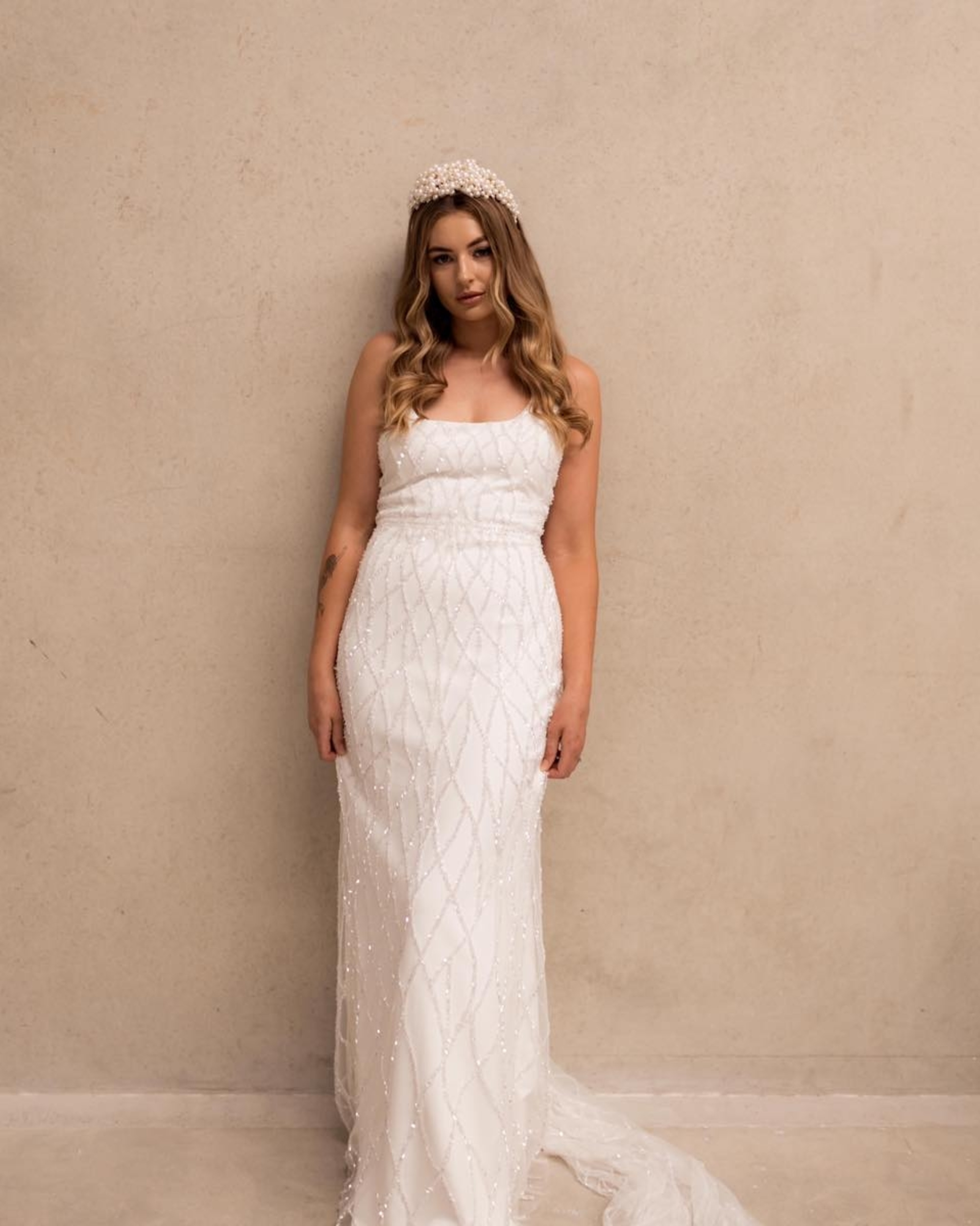 Bride in a beaded white fitted wedding dress and pearl headpiece standing against a neutral wall