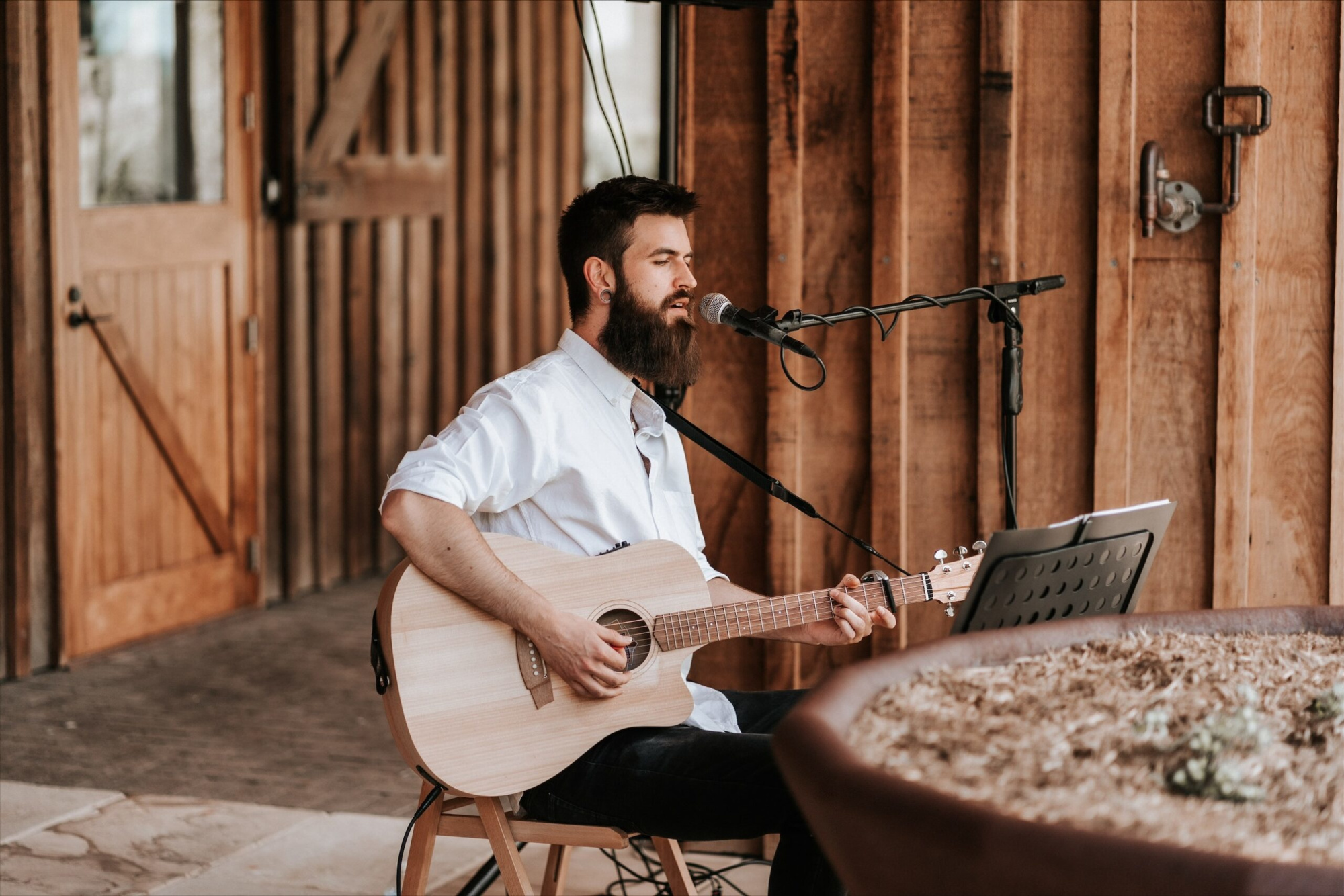 Bearded acoustic guitarist singing into a microphone at a rustic wooden wedding venue.