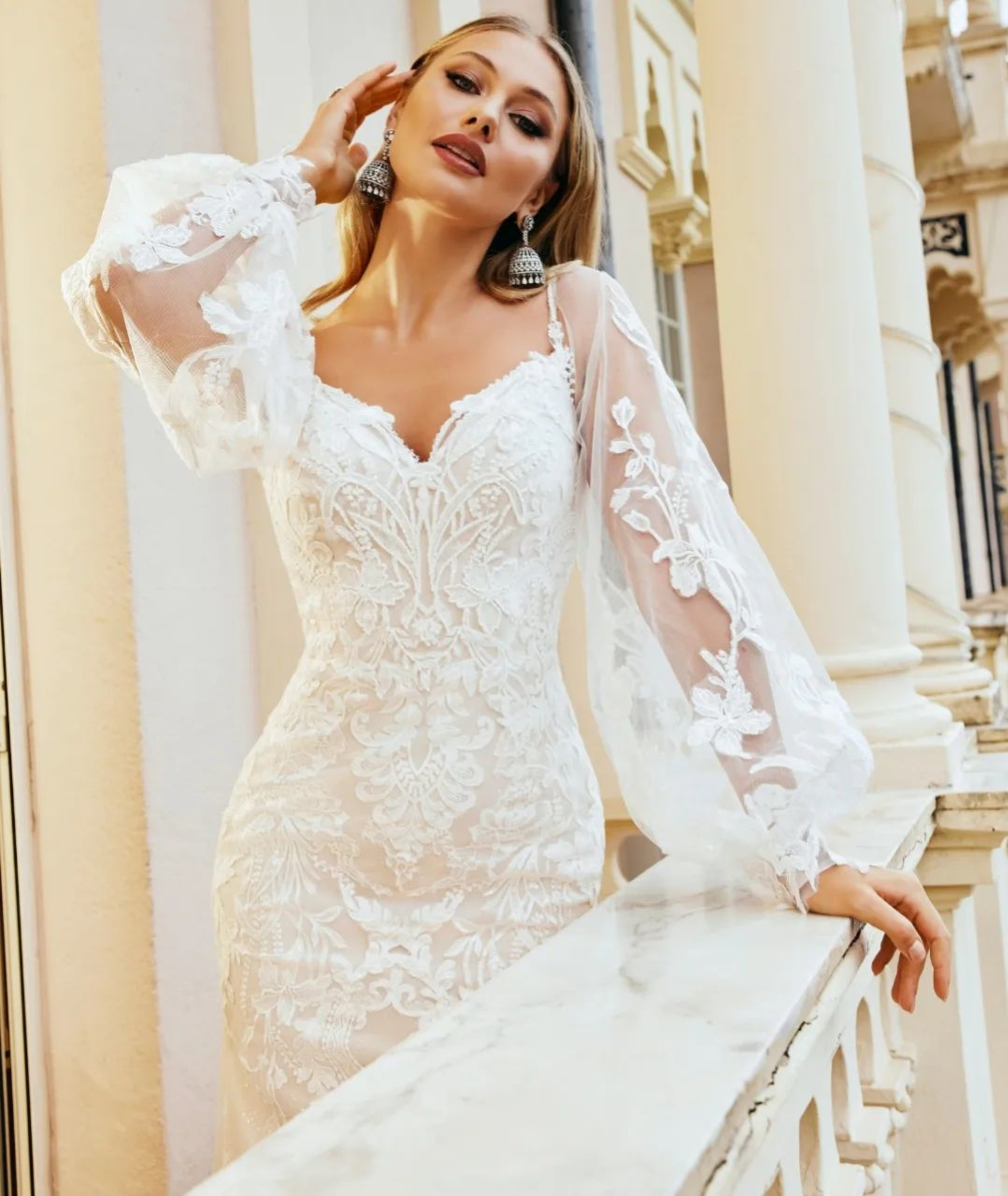 Bride in a fitted lace wedding gown with sheer floral sleeves posing on an elegant balcony.