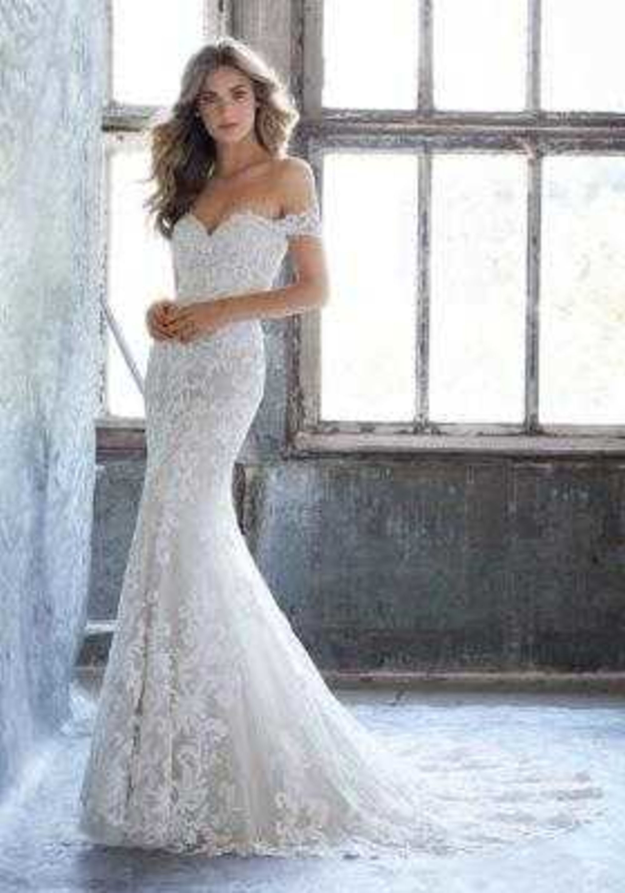 Bride in an off-the-shoulder lace mermaid wedding dress standing by a large rustic window.