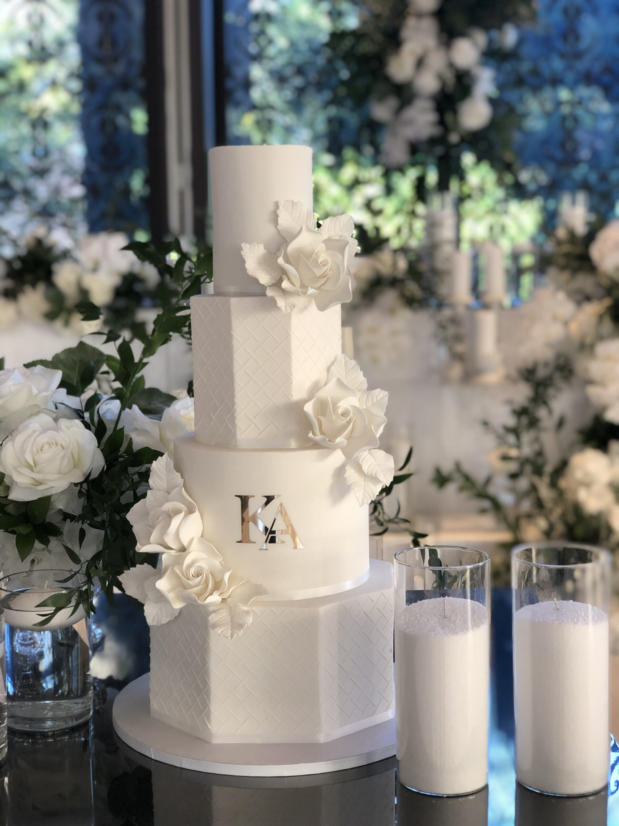 Tall white tiered wedding cake with sugar roses and monogram, surrounded by candles and floral decor.