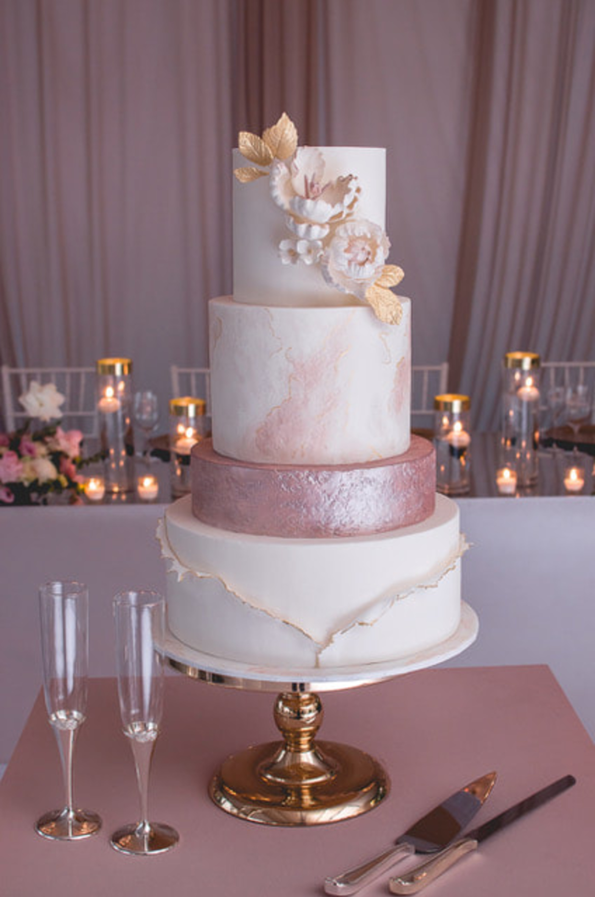 Tall four-tier blush and white wedding cake with gold accents and sugar flowers on a gold stand at a candlelit reception table.