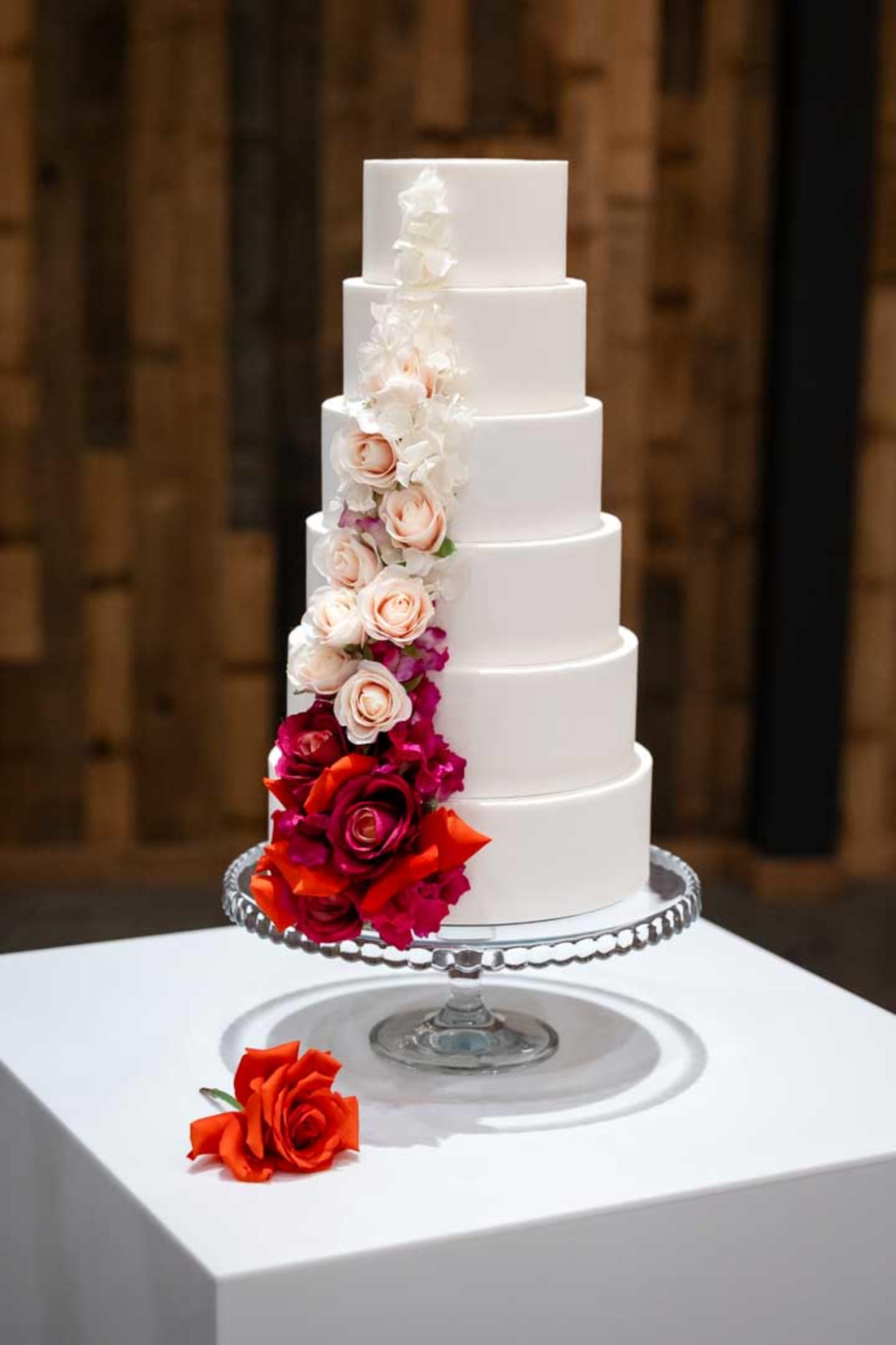 Tall white tiered wedding cake decorated with cascading vibrant roses on a glass cake stand.