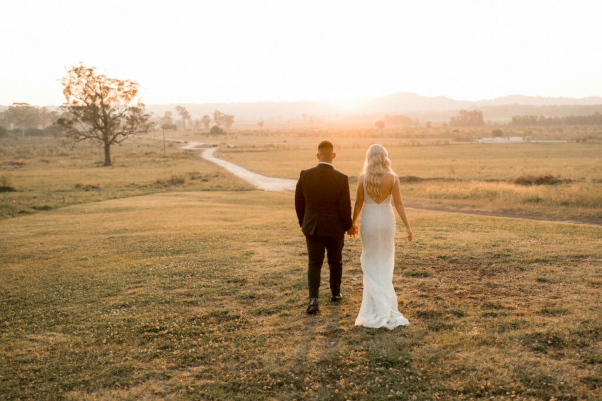 Bride and groom walk hand in hand across a sunlit field at sunset on their wedding day.