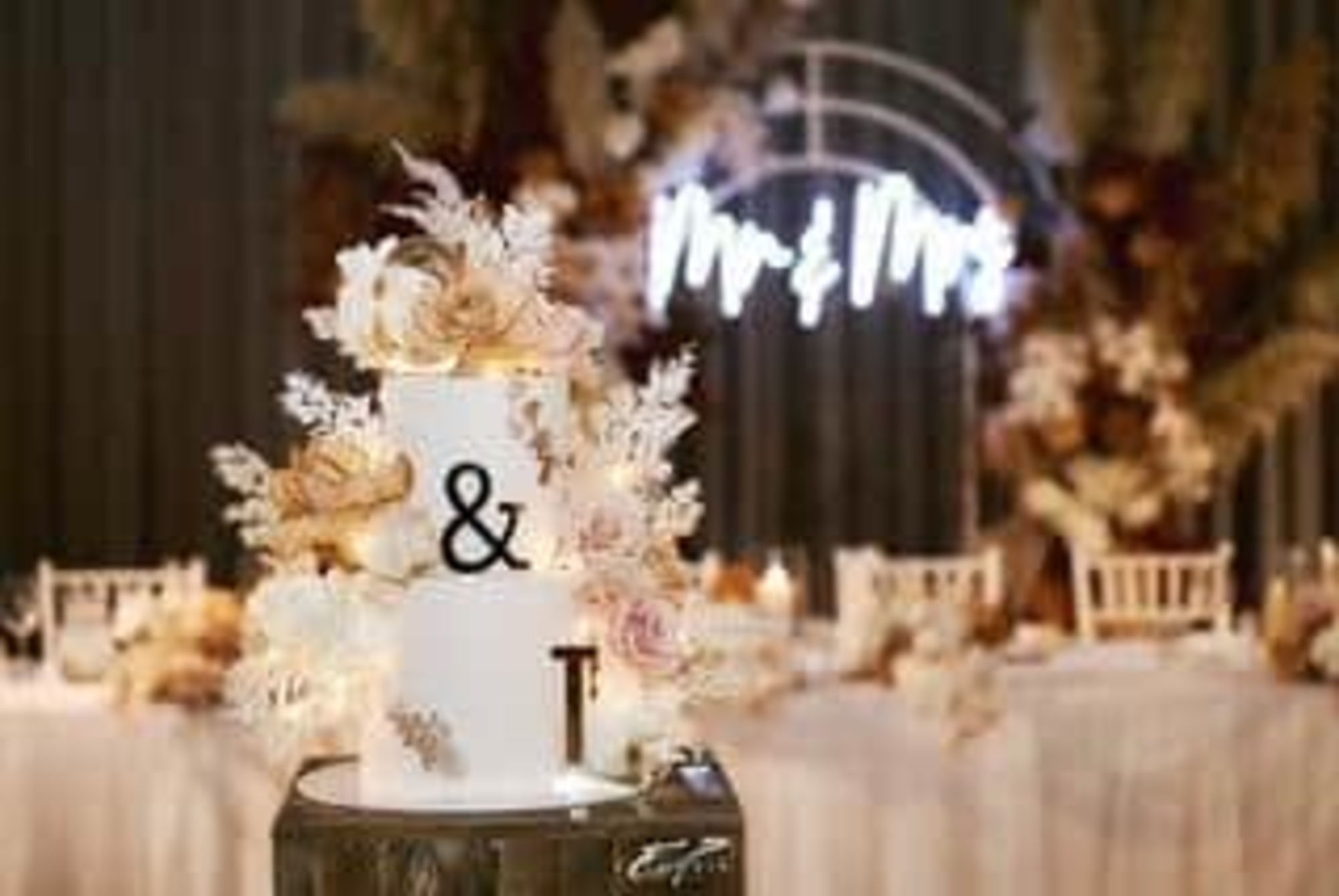 Elegant white tiered wedding cake with neutral florals in front of a softly lit Mr and Mrs reception backdrop.
