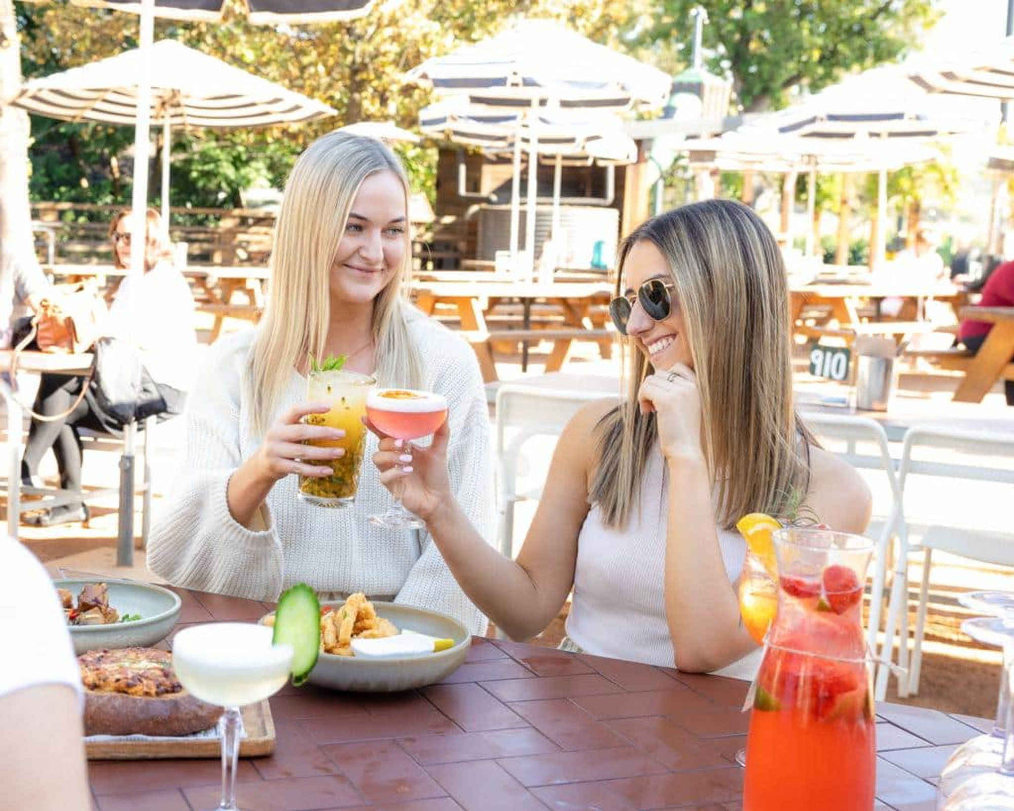 Two women enjoy colorful cocktails at an outdoor patio table with food and drinks.