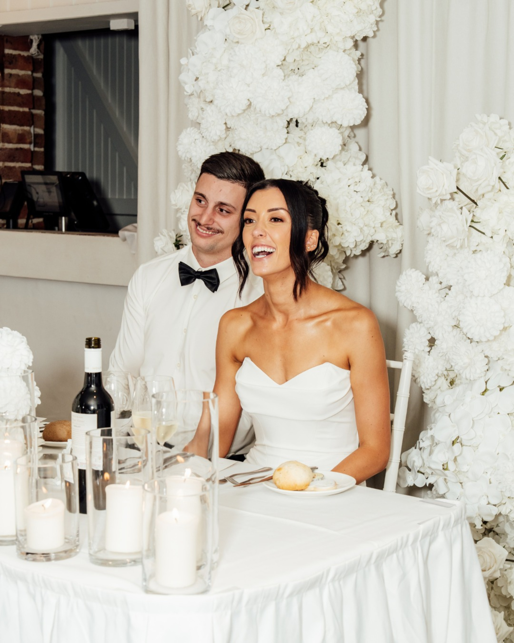 Bride and groom sit smiling at a white sweetheart table with candles and a lush white floral backdrop.