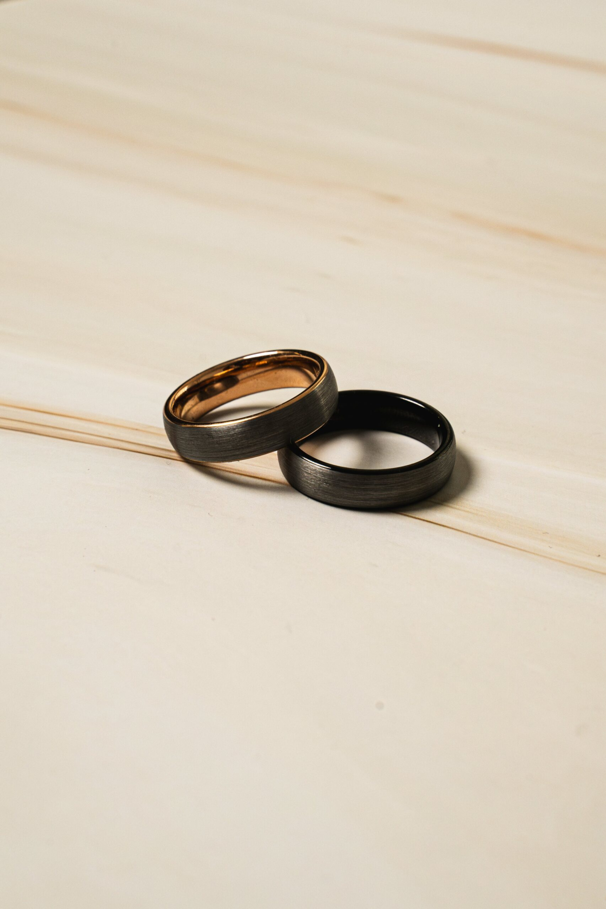 Two modern metal wedding bands rest overlapped on a light wooden surface.