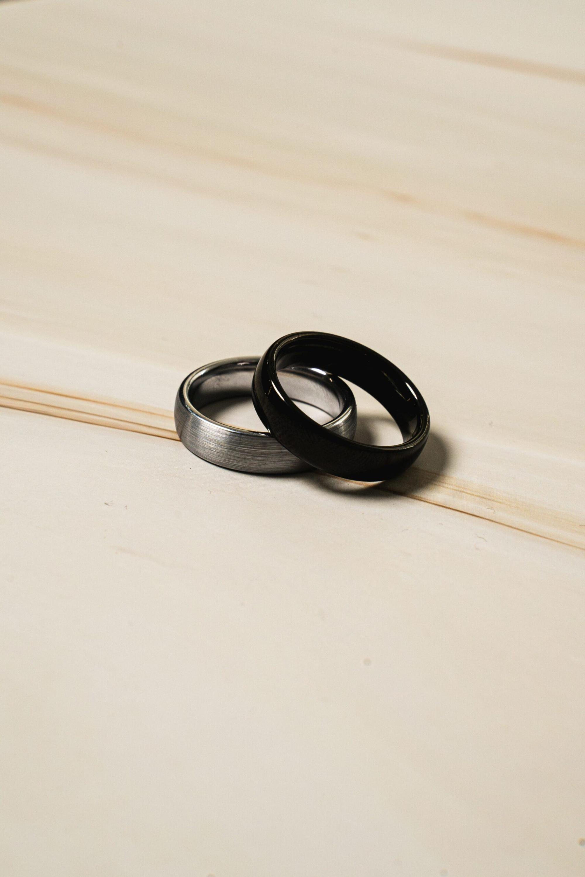 Two modern metal wedding bands, one black and one silver, rest crossed on a light wood surface.
