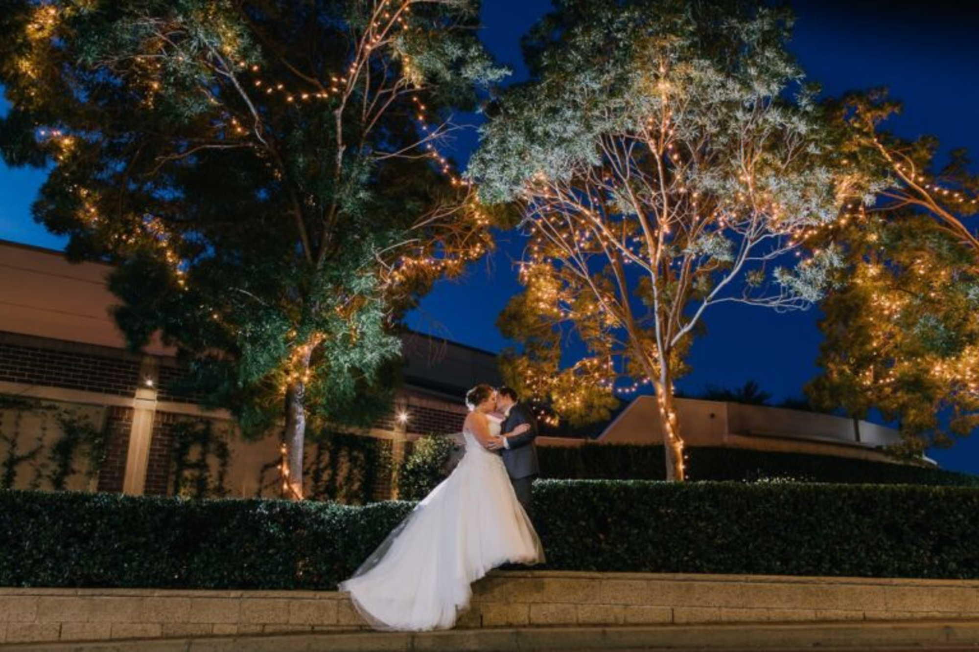 Bride and groom share a romantic embrace under twinkling string lights in an outdoor garden at night.