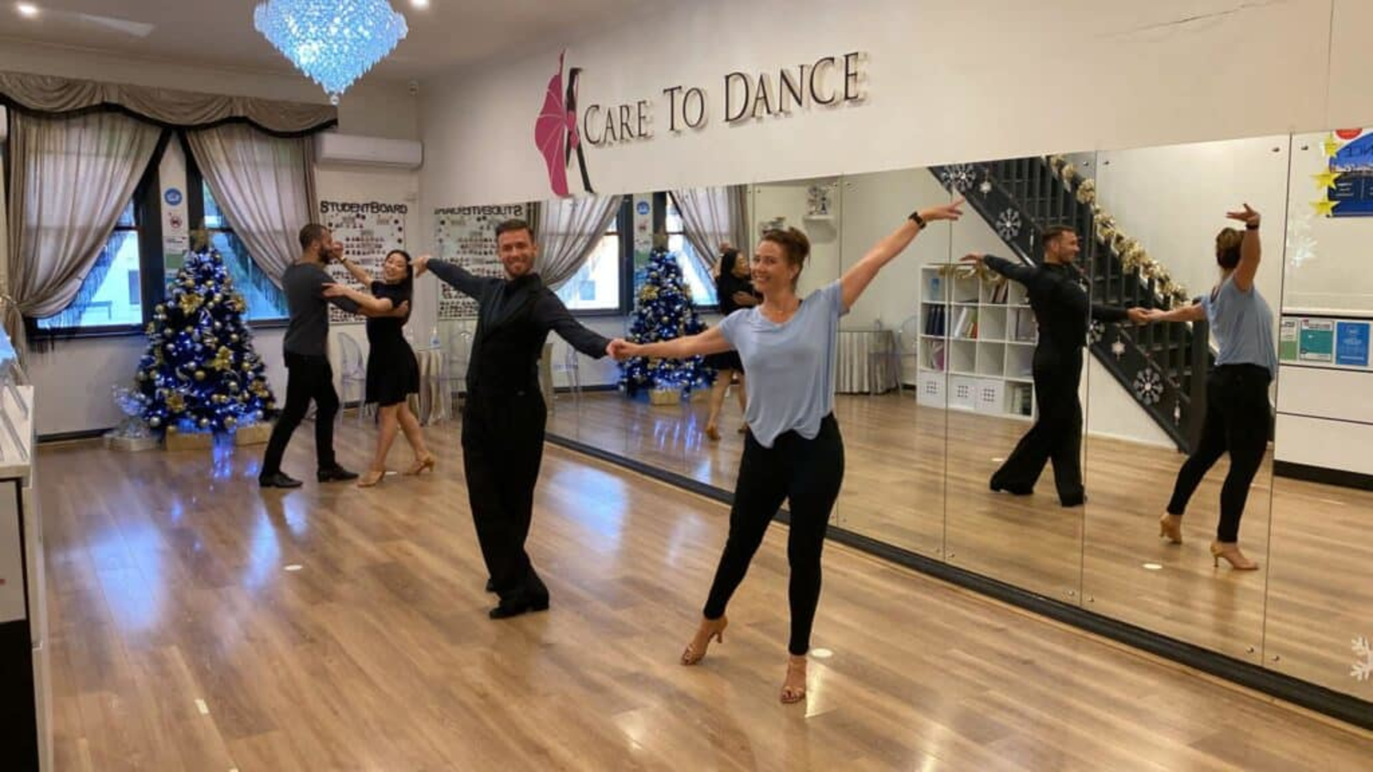 Couples practice ballroom steps with instructors in a bright dance studio decorated for the holidays.