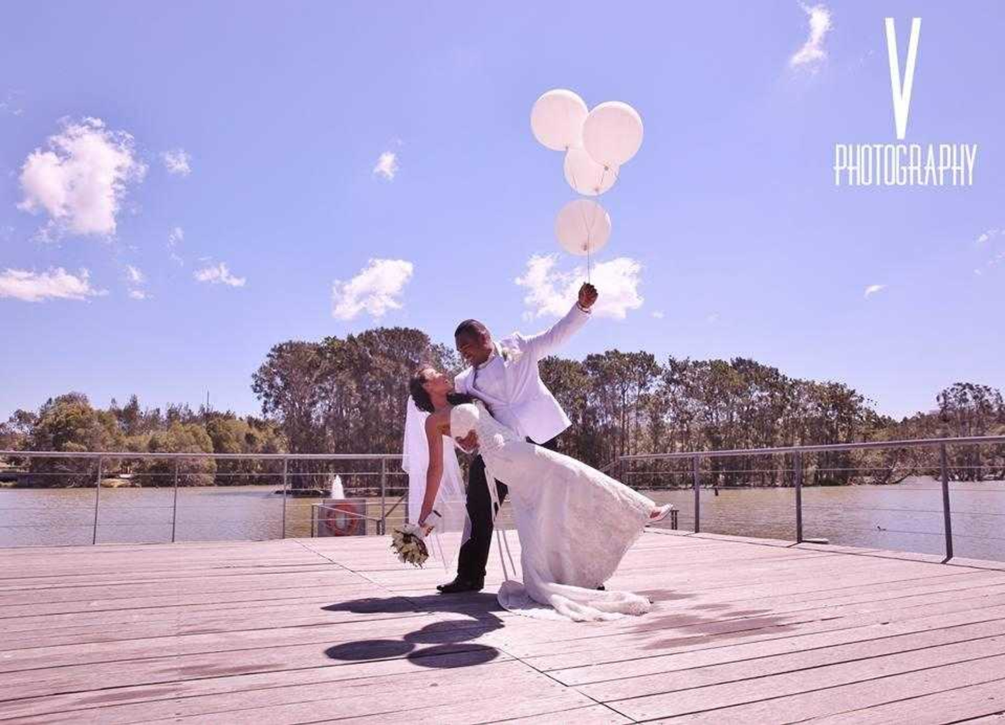 Bride and groom share a romantic dip on a lakeside deck holding white balloons under a bright sky.