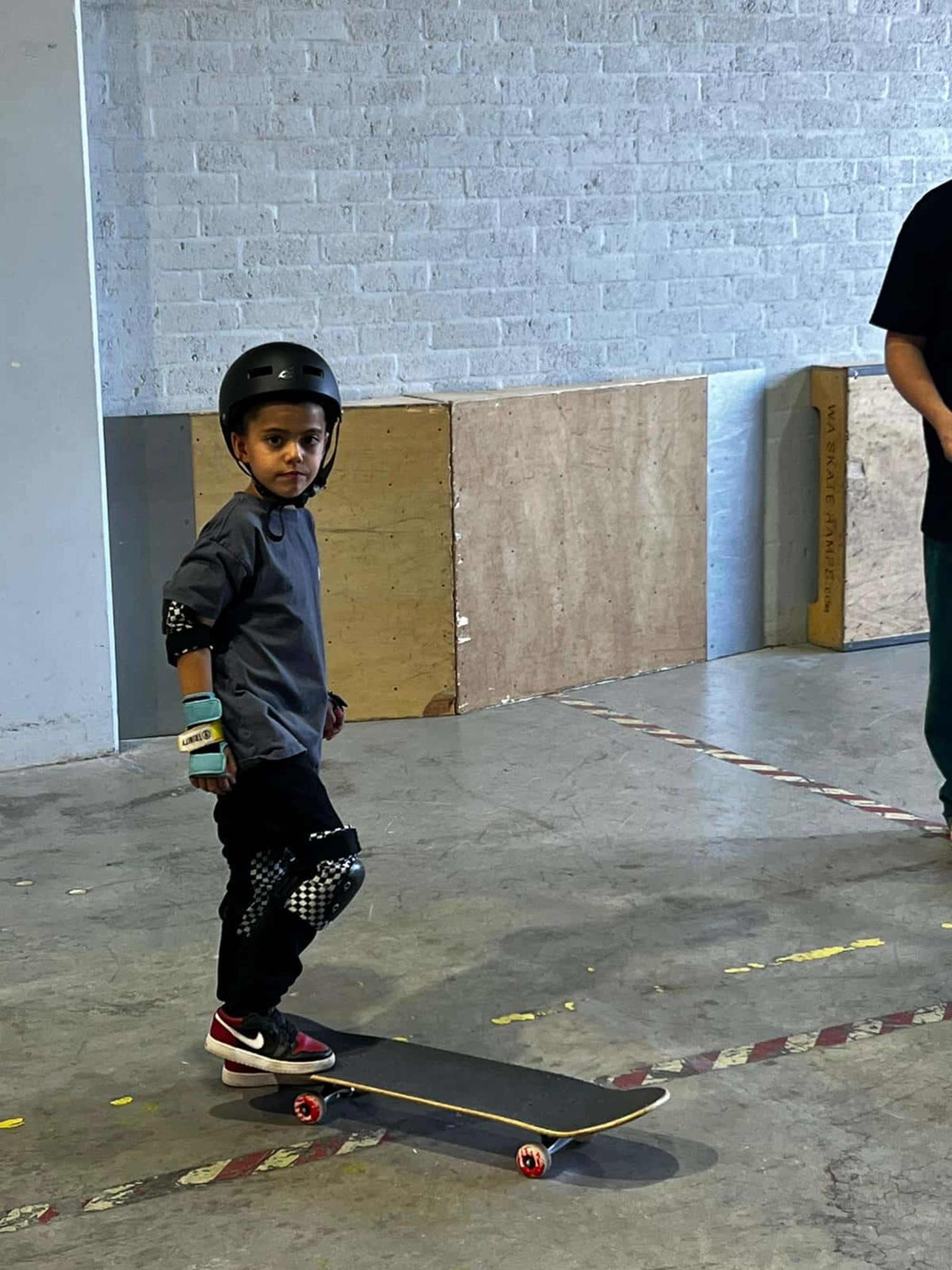 Child wearing safety gear standing on a skateboard in an indoor skatepark.