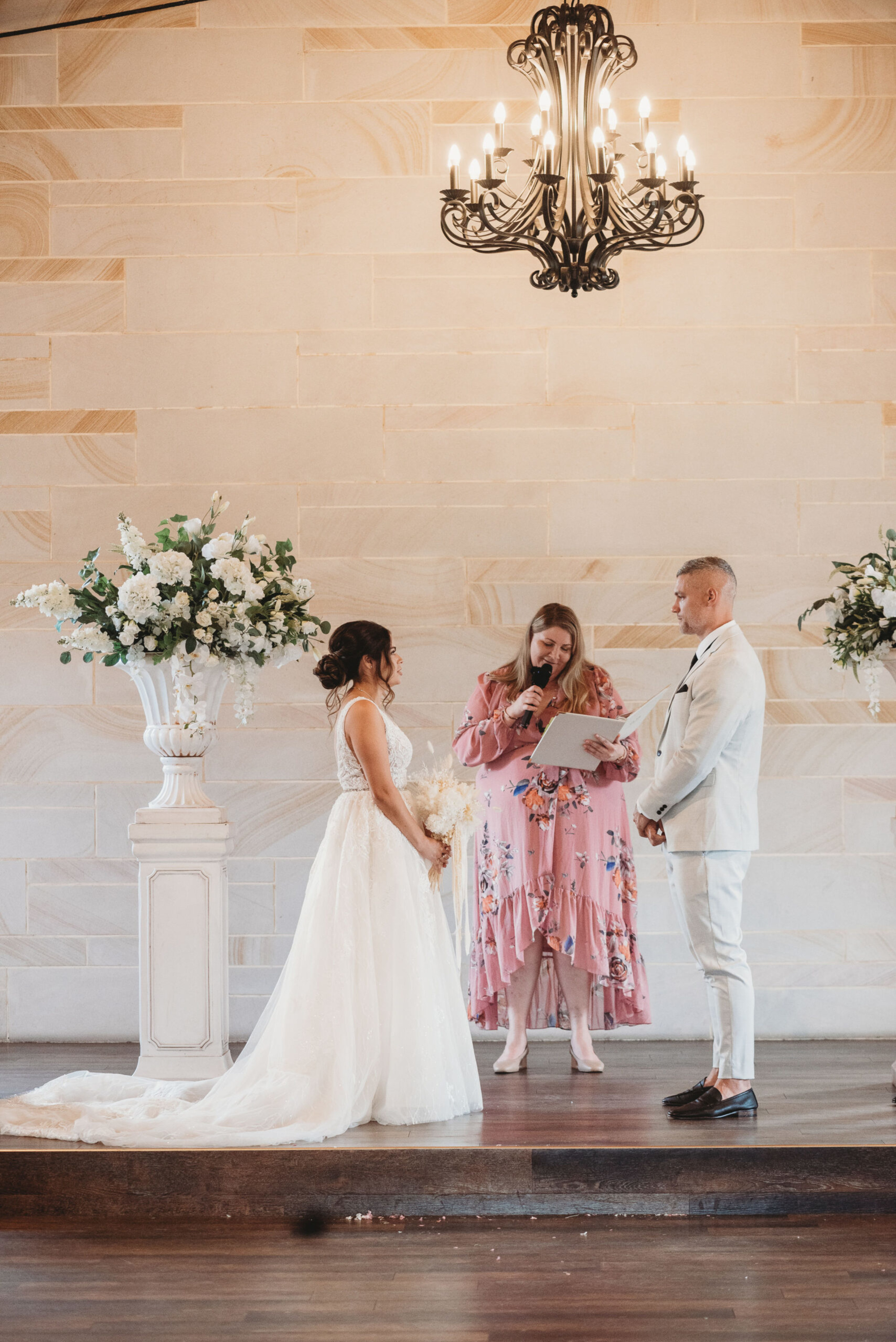 Bride and groom stand with officiant under a chandelier during an indoor wedding ceremony with white floral arrangements.
