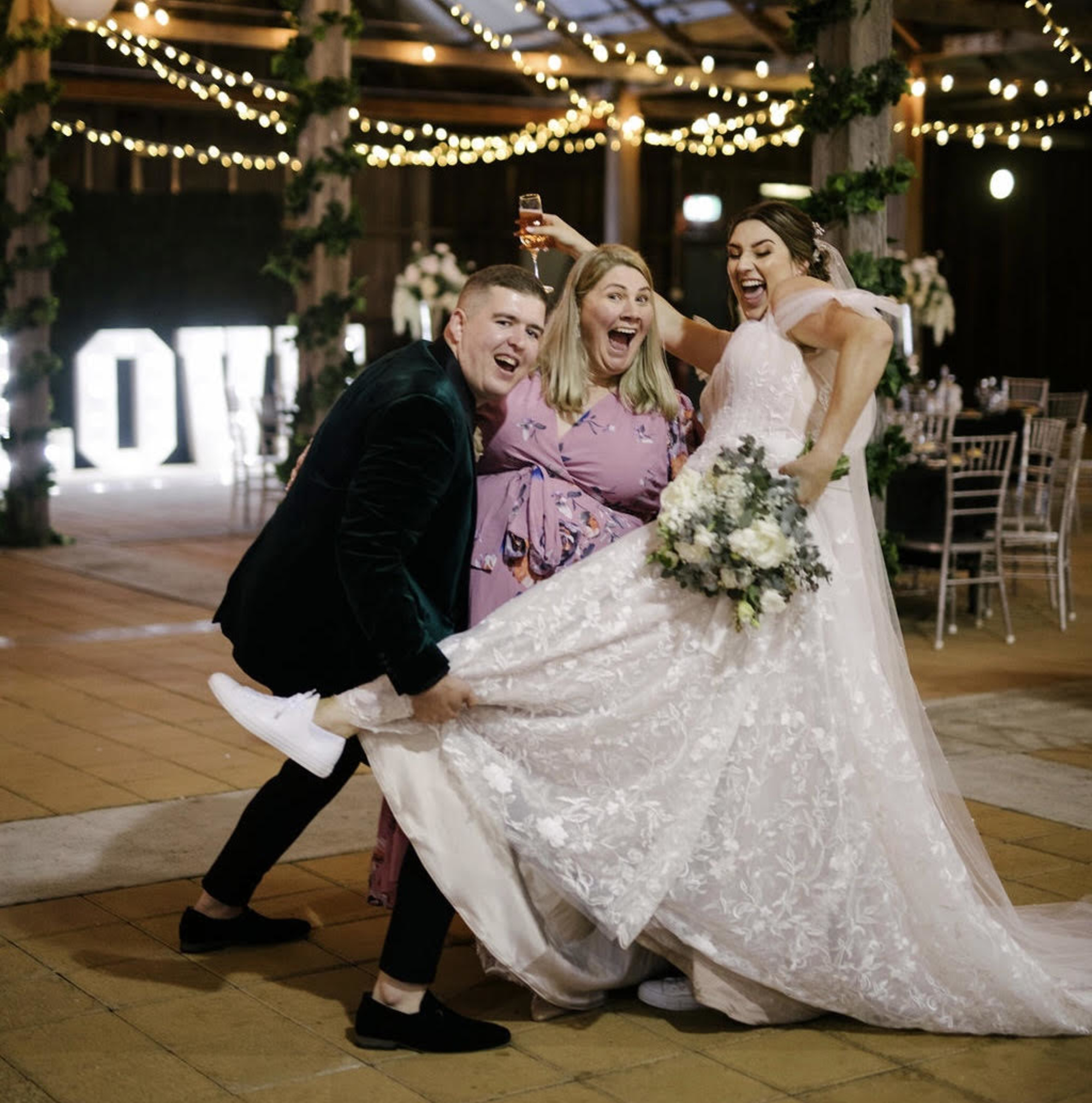 Joyful bride and groom pose playfully with a guest under string lights at an indoor wedding reception.