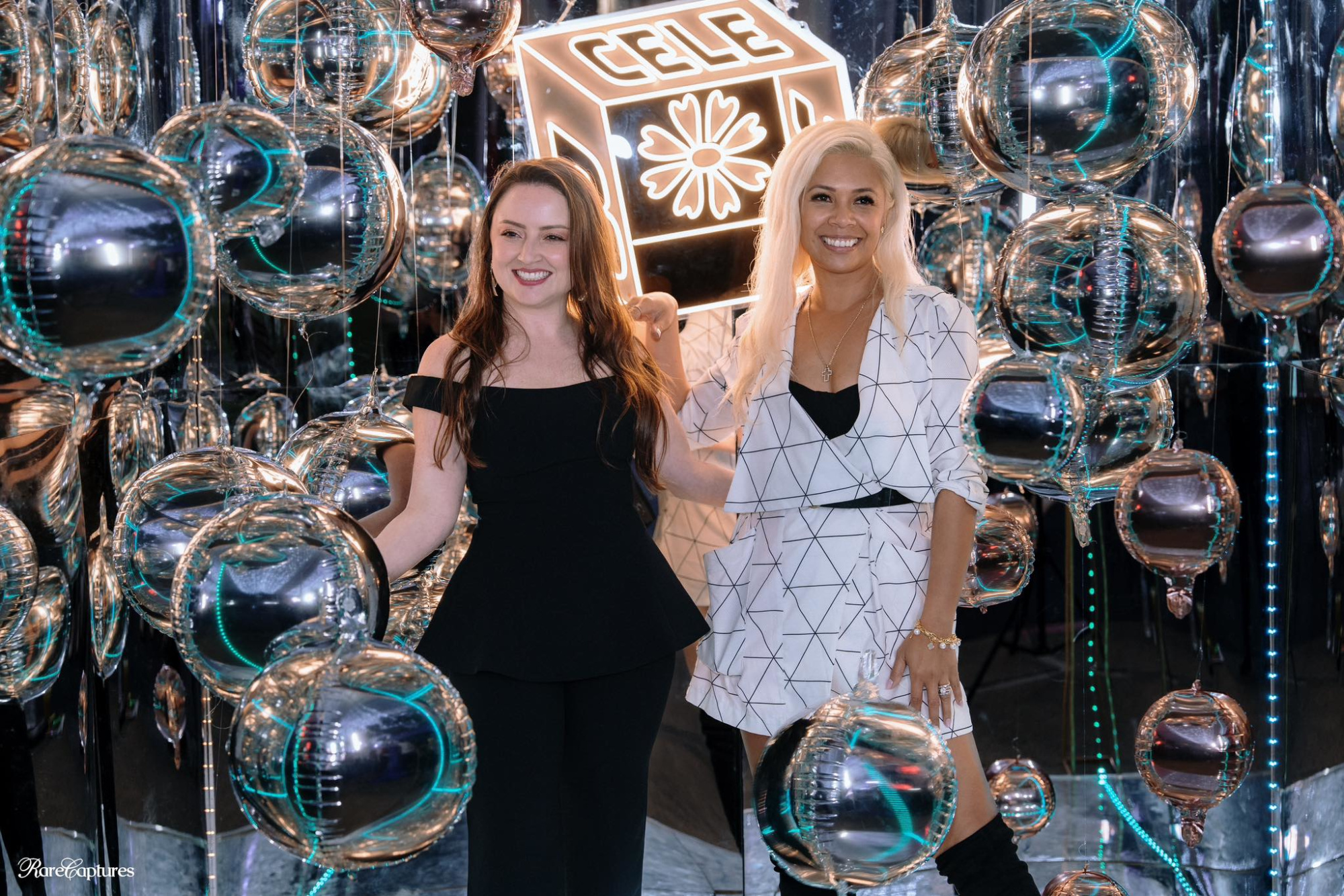 Two women pose in front of a neon wedding sign surrounded by reflective metallic balloon decor.