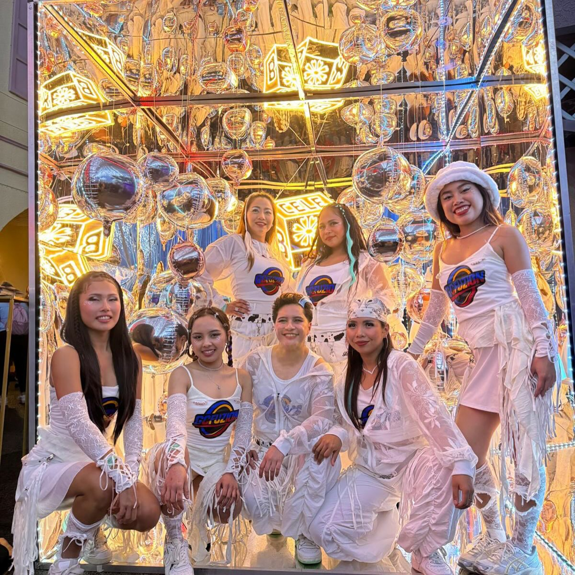 Group of performers in coordinated white outfits posing in a mirrored, glowing installation with metallic balloons.