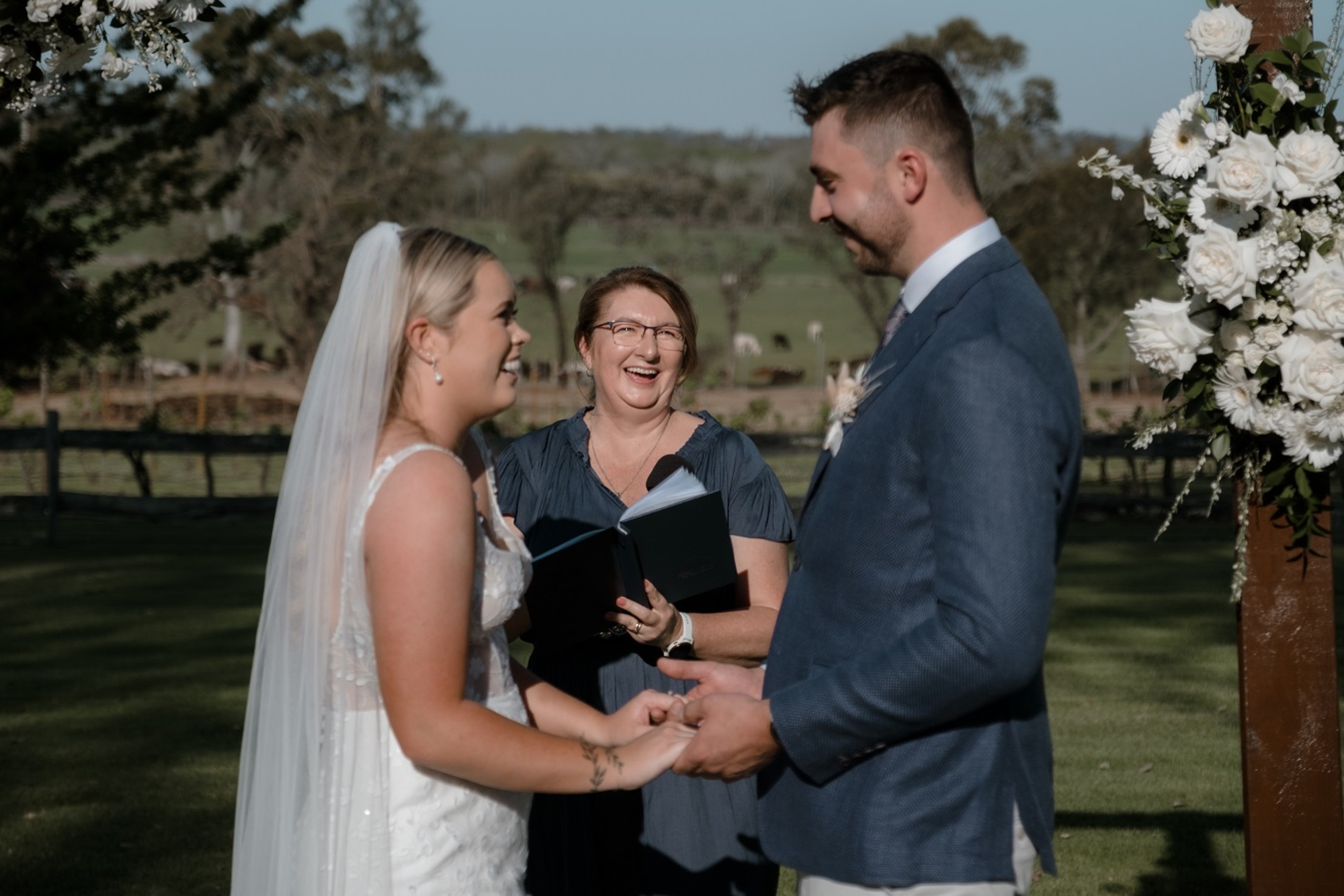 Couple holding hands and smiling during an outdoor wedding ceremony with a celebrant and floral arch.