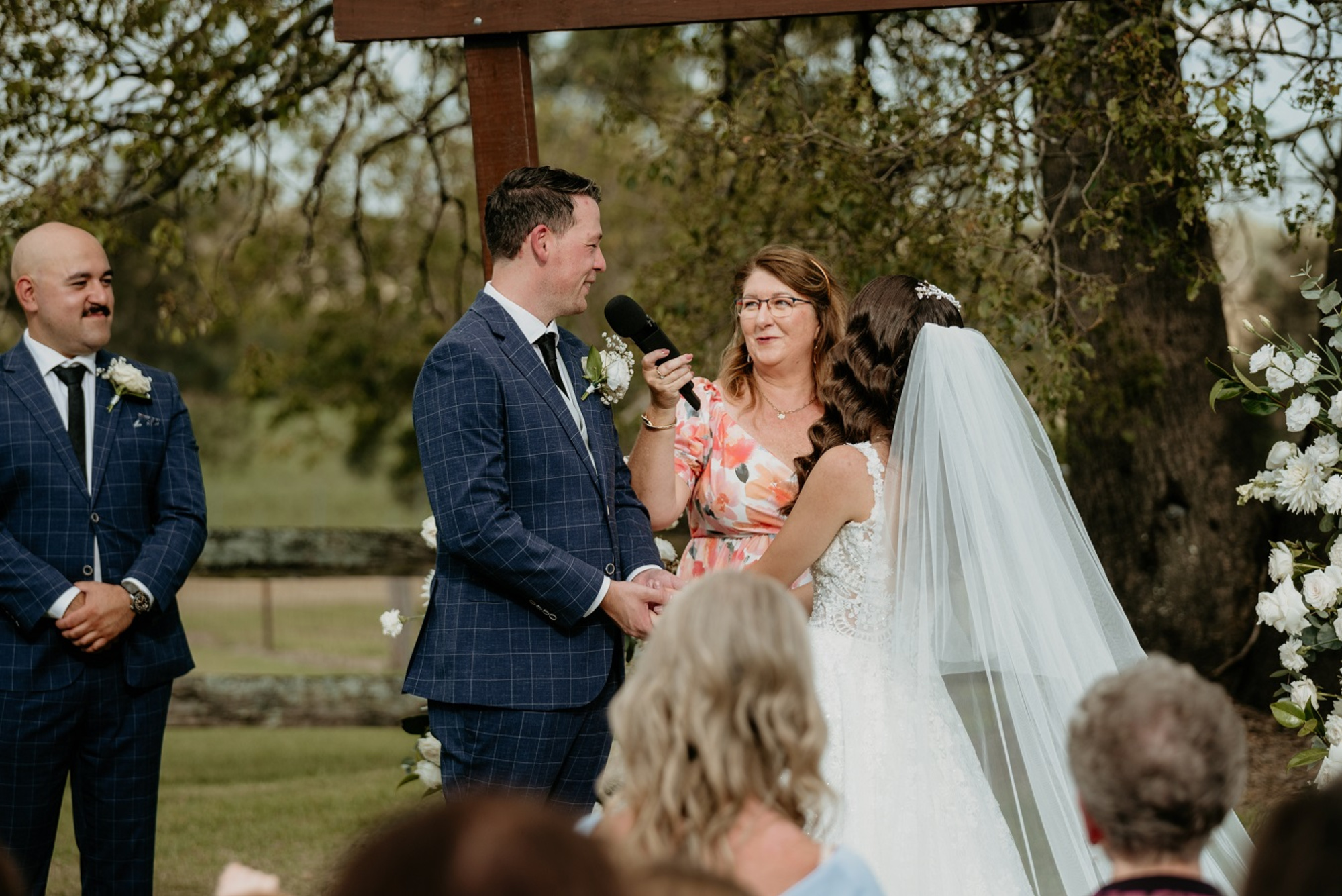 Outdoor wedding ceremony with a couple holding hands as the celebrant speaks into a microphone under a rustic wooden arch.