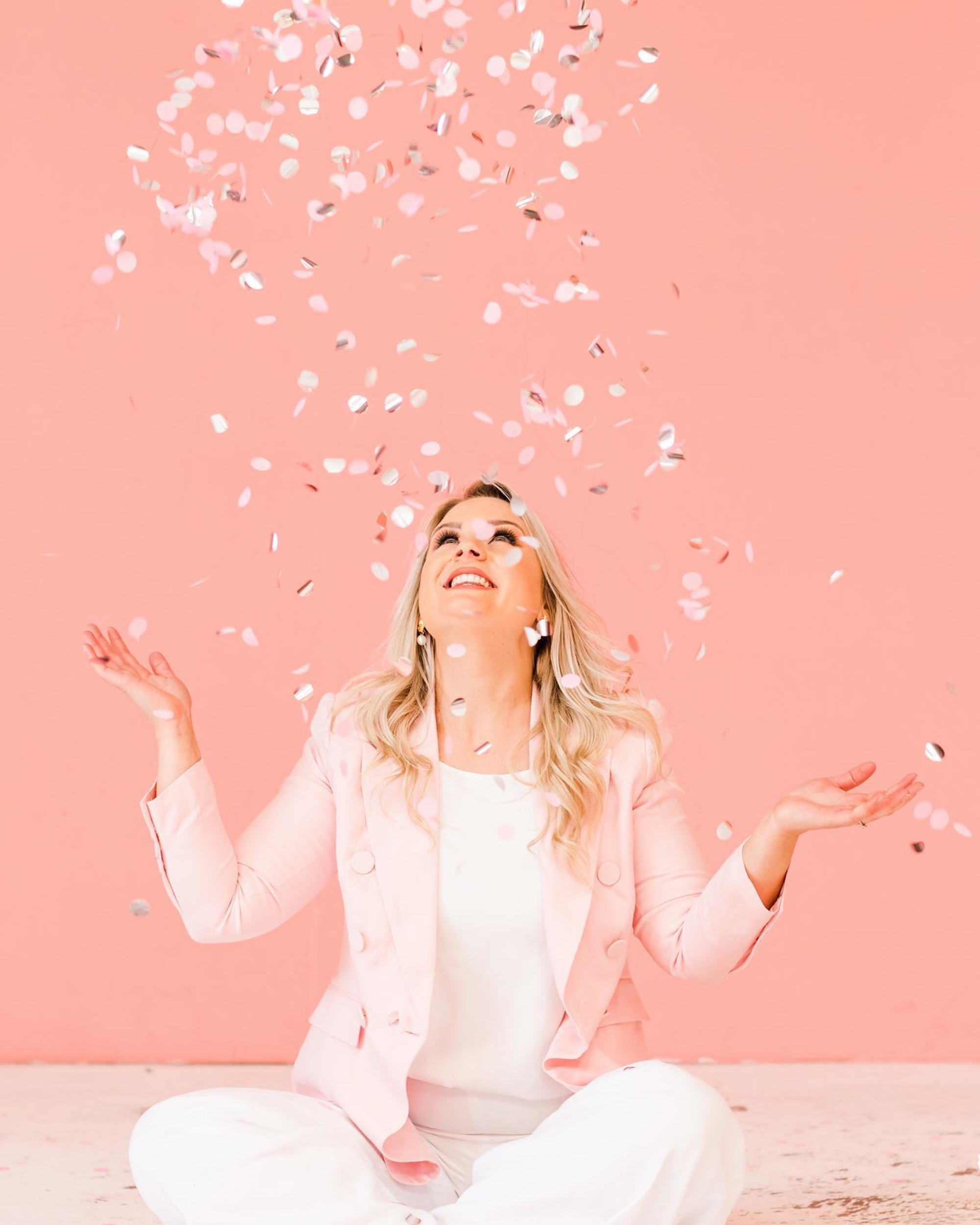 Smiling woman in light pink blazer sits against a pink backdrop tossing confetti in the air.