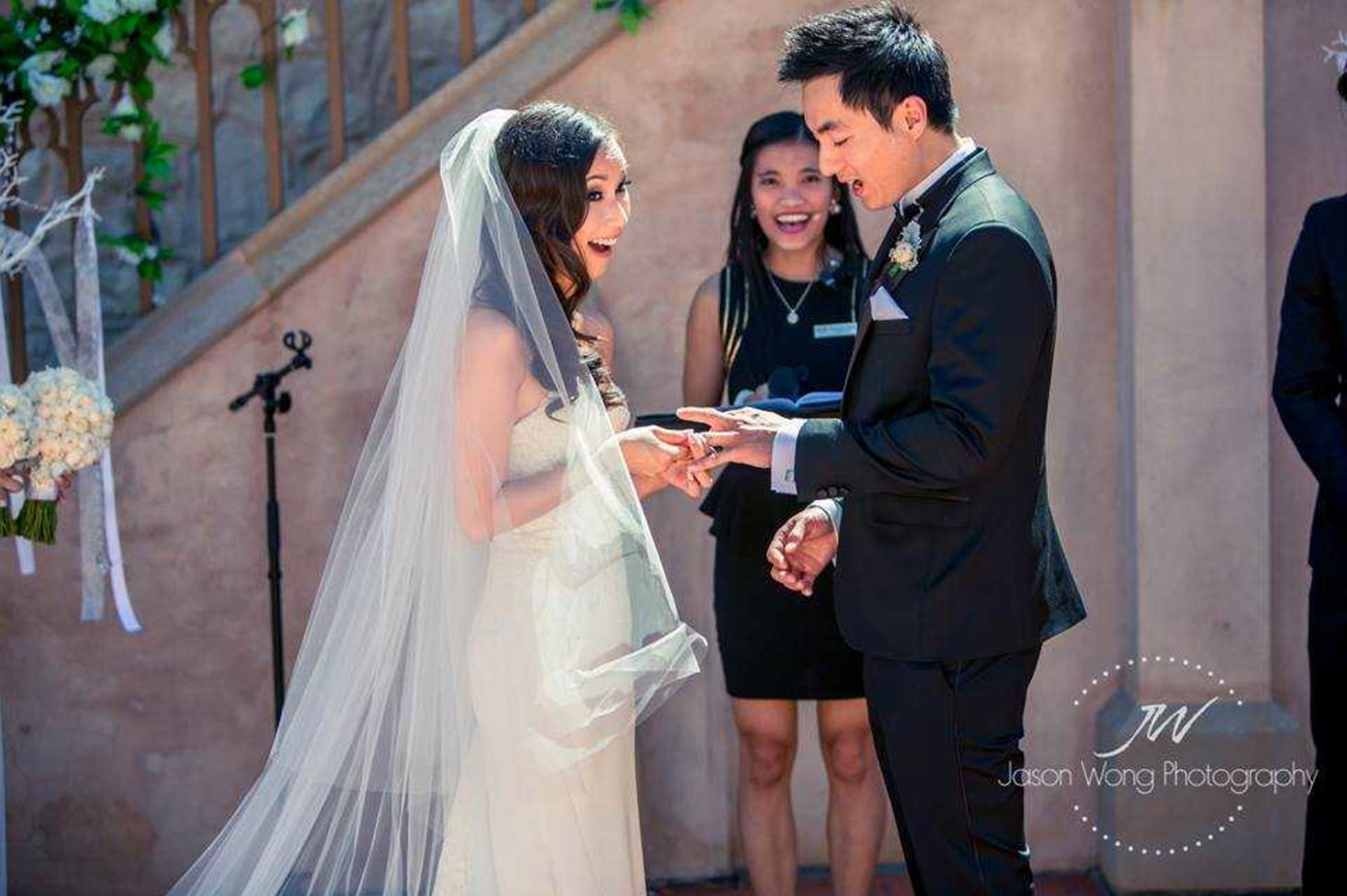 Bride and groom joyfully exchange rings during an outdoor wedding ceremony with an officiant smiling behind them.