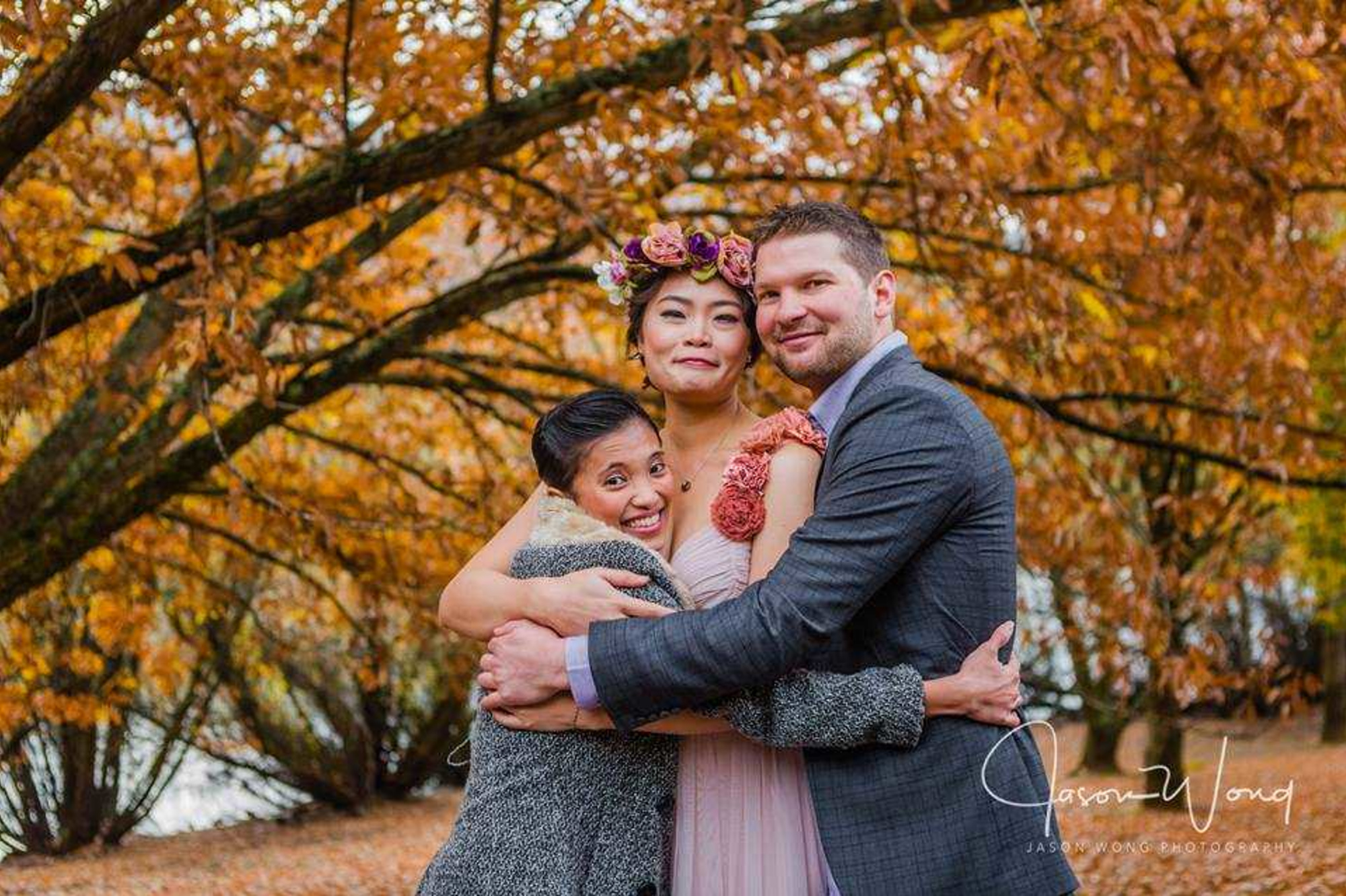 Smiling wedding couple and guest hugging under golden autumn trees in a park.