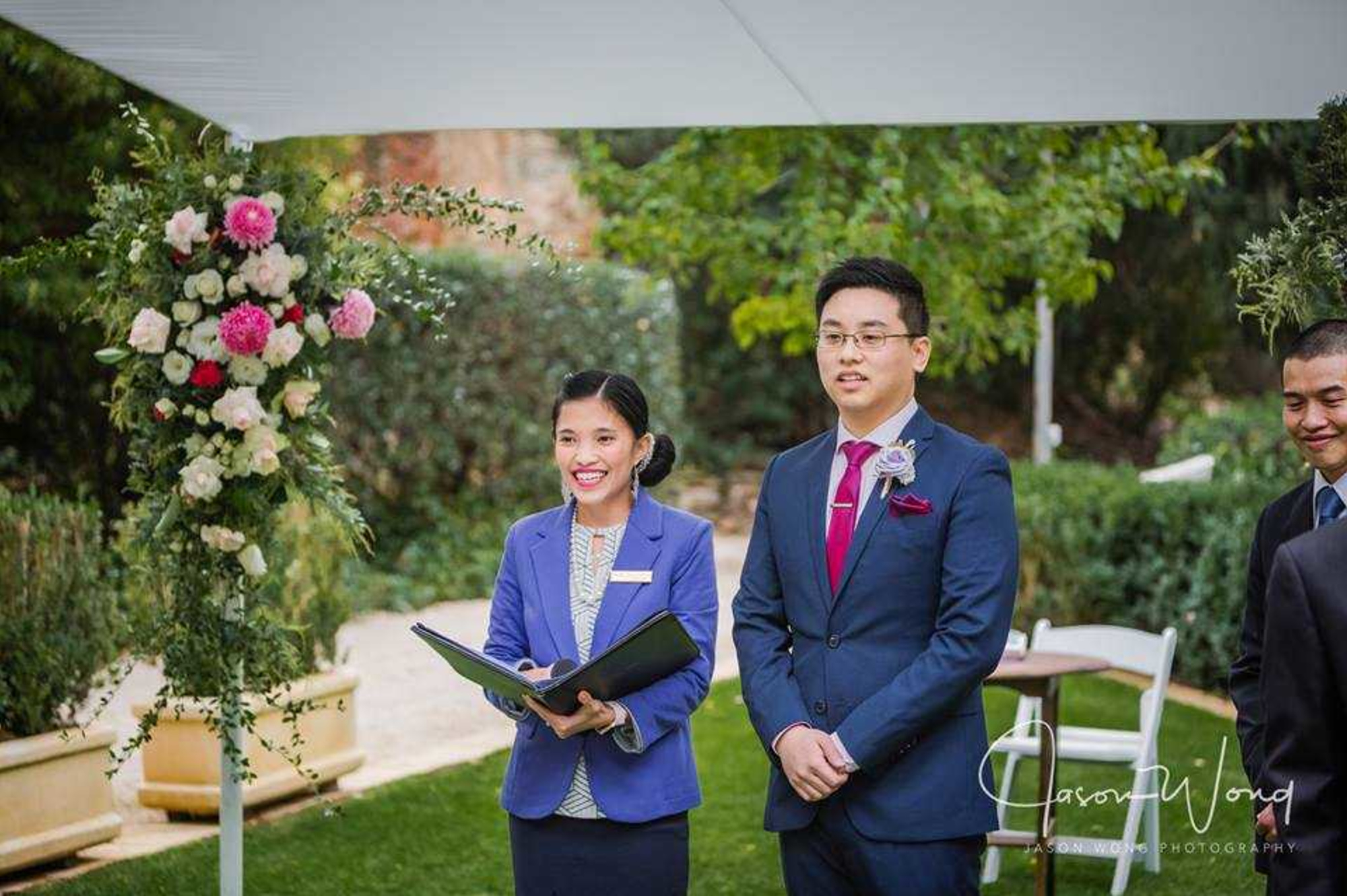 Groom and officiant stand at an outdoor garden wedding ceremony beside a floral arrangement.