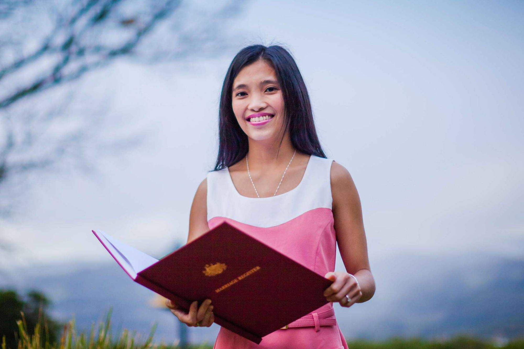 Smiling wedding celebrant holds an open marriage registry folder outdoors with mountains in the background.
