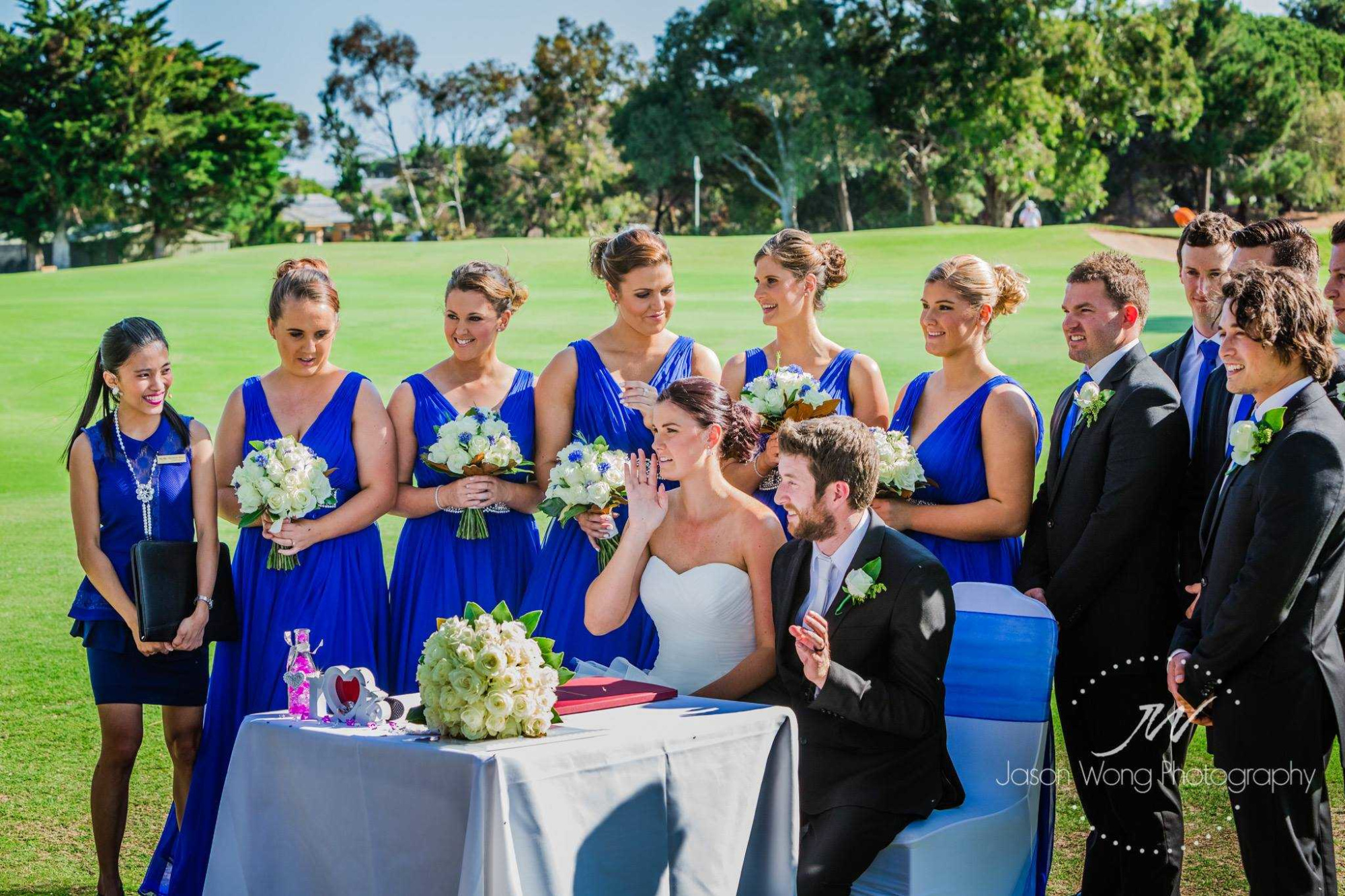 Outdoor wedding ceremony with bride, groom, and bridal party in blue dresses gathered around a signing table on a lawn.