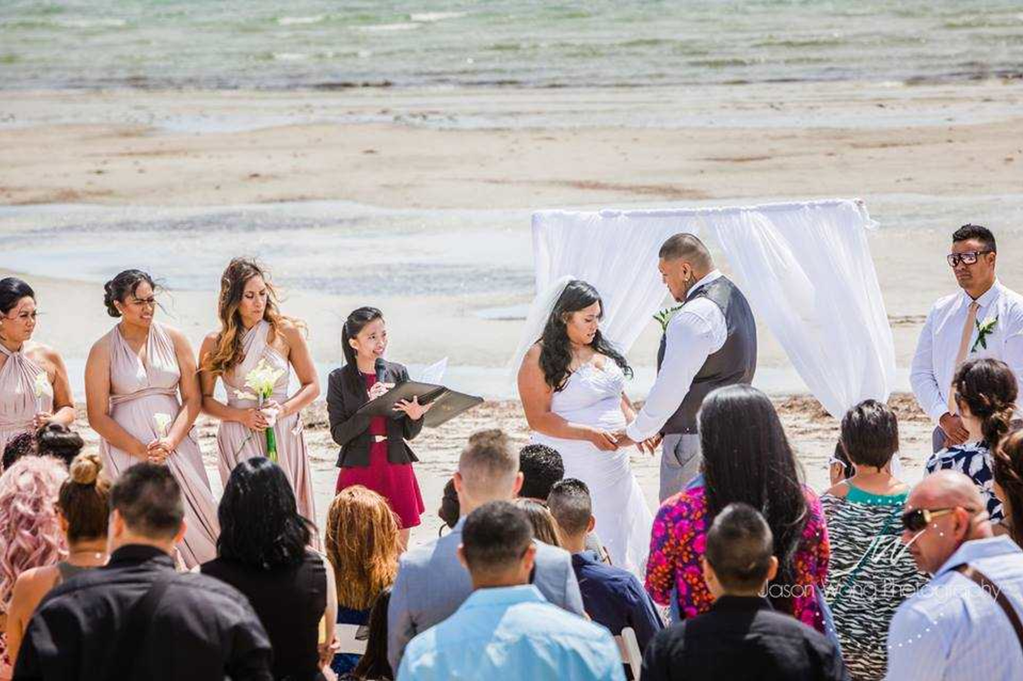 Couple exchange vows beneath a white draped arch during a beach wedding ceremony with guests and bridal party watching.