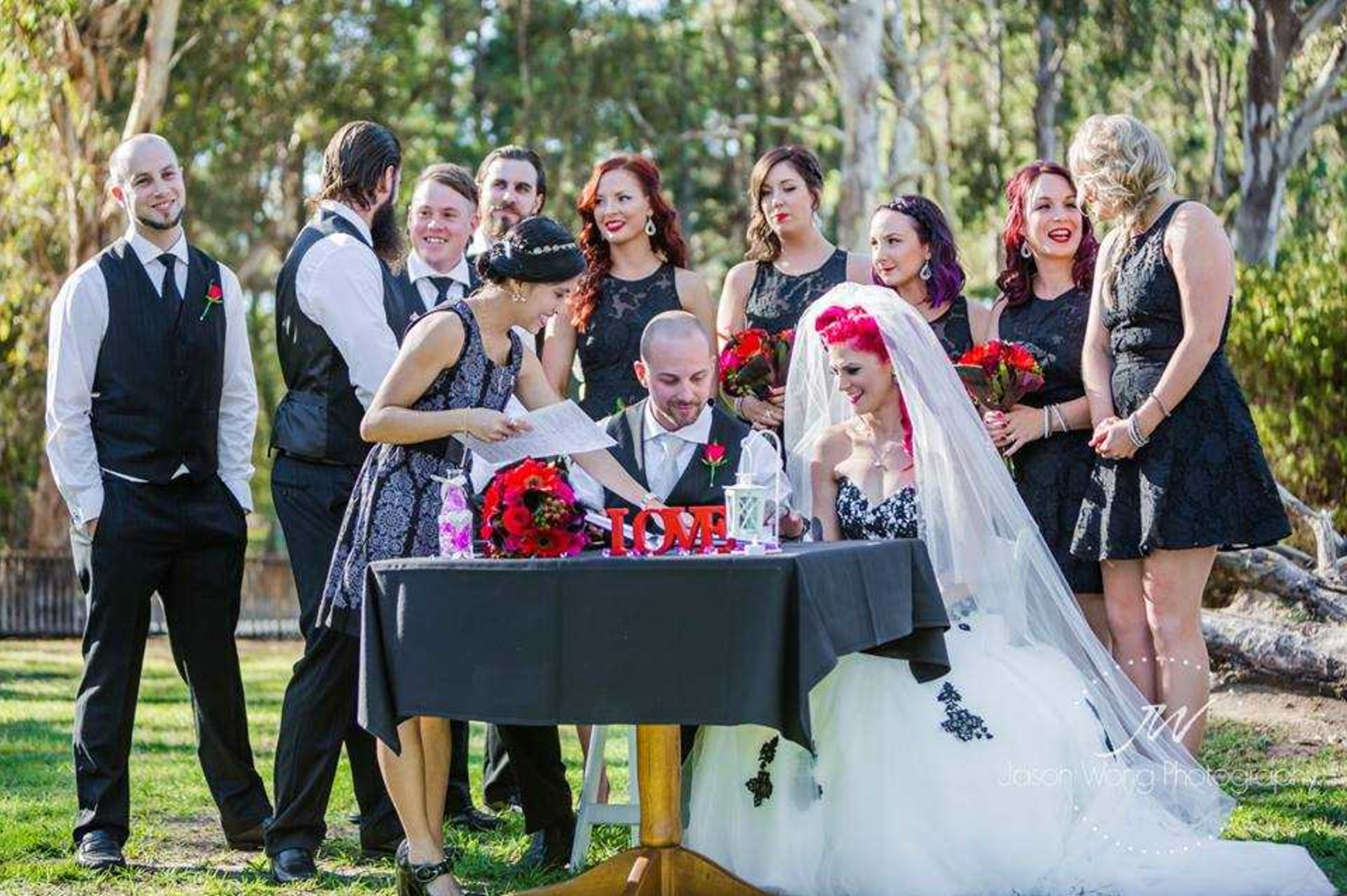 Bride and groom sign their marriage certificate outdoors surrounded by joyful bridesmaids and groomsmen.