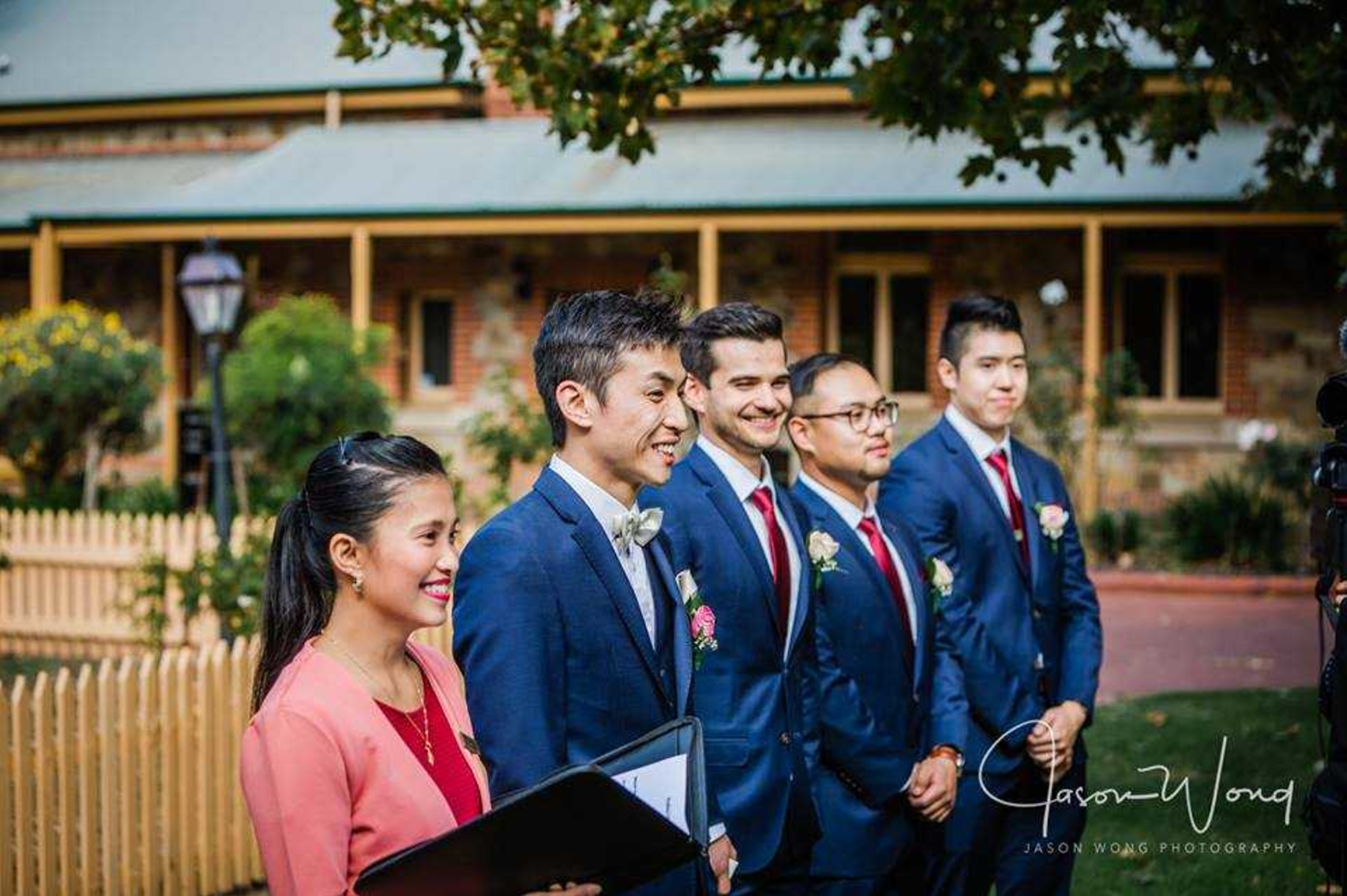 Groom and groomsmen in navy suits smile during an outdoor wedding ceremony at a garden venue.