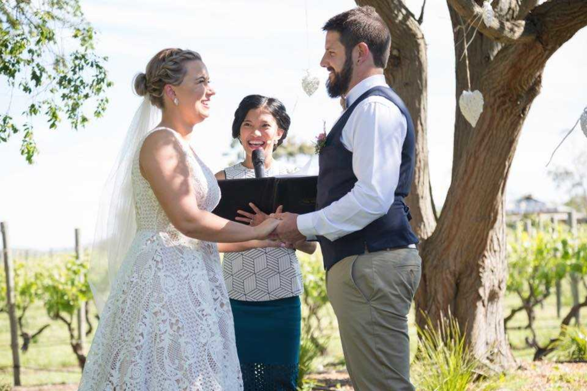 Bride and groom hold hands during an outdoor vineyard ceremony led by a smiling officiant under a tree.