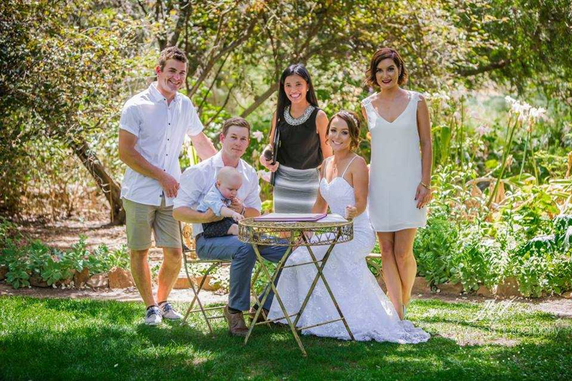 Bride and groom with family and officiant pose by a signing table in a sunny garden wedding setting.