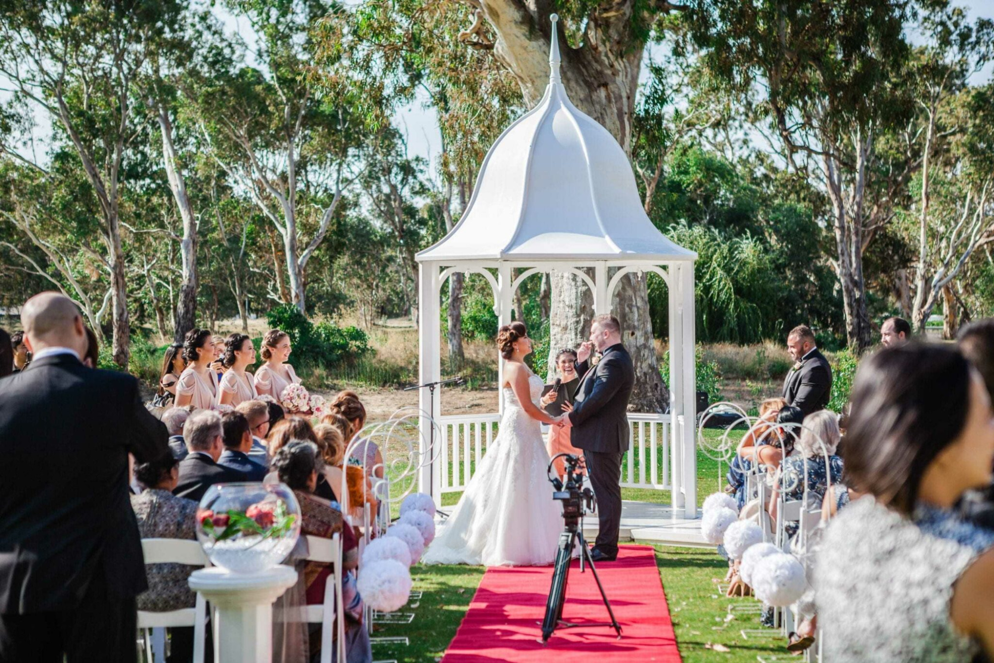 Outdoor wedding ceremony under a white gazebo with a red carpet aisle and guests seated on either side.