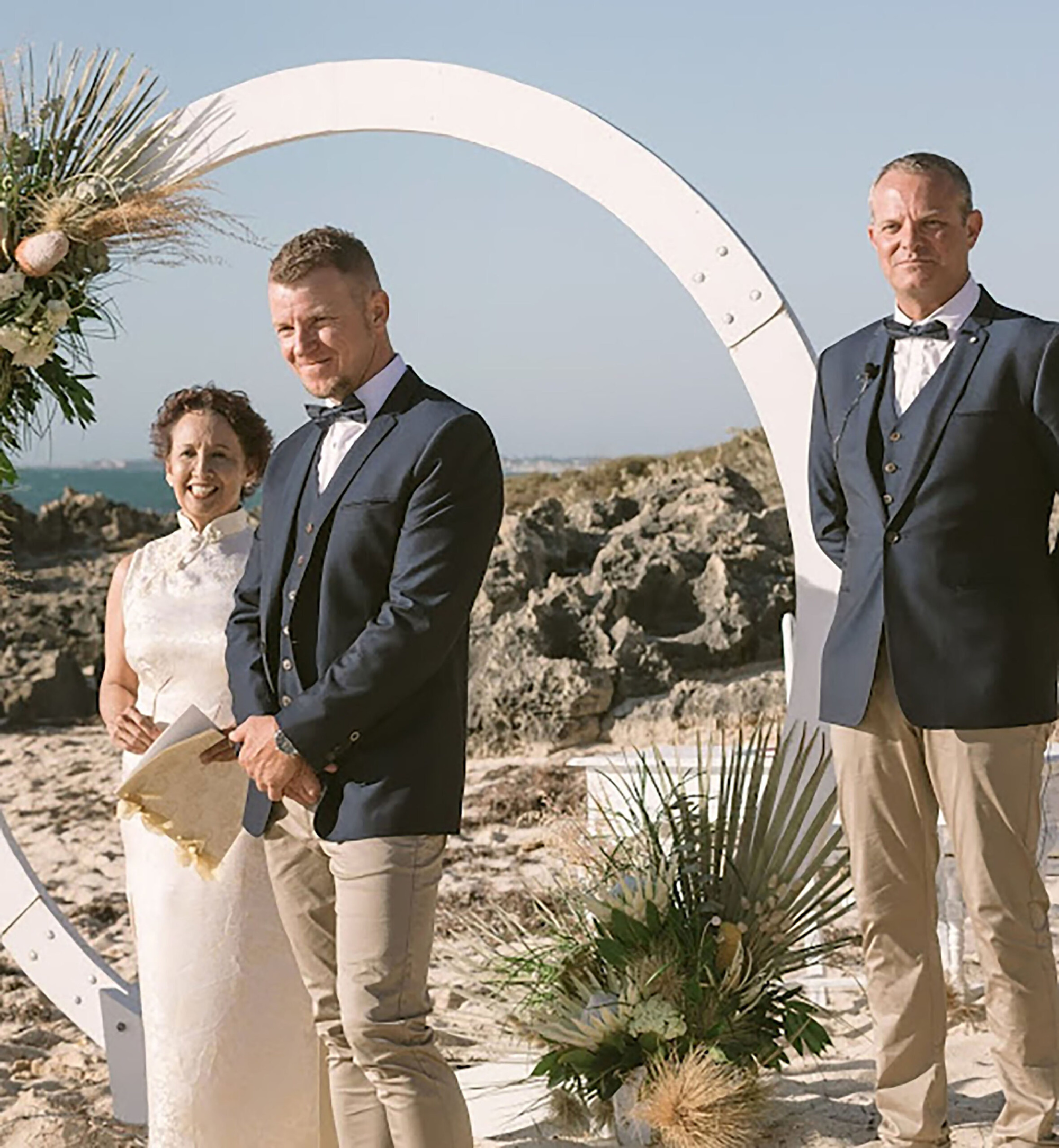 Groom and officiant stand under a modern circular arch at a beach wedding ceremony with coastal rock backdrop.