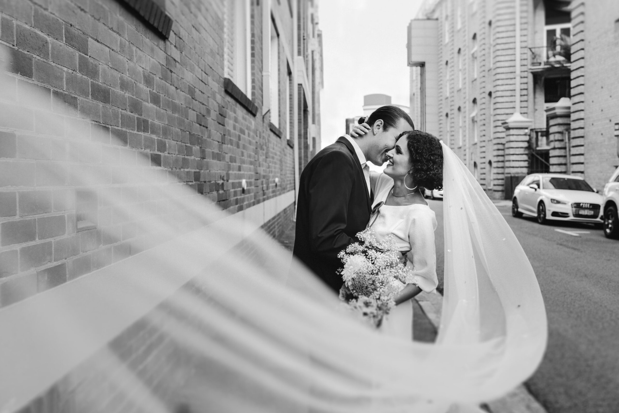 Black and white photo of a couple embracing on a city street with the bride’s veil flowing in the foreground.